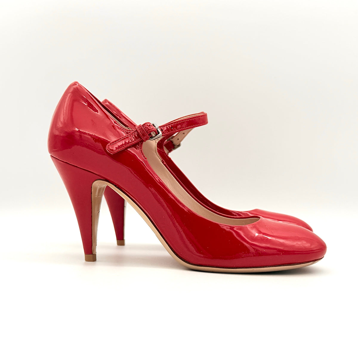 A pair of red patent leather Miu Miu Mary Jane heels with adjustable buckle straps and high heels, displayed against a white background.