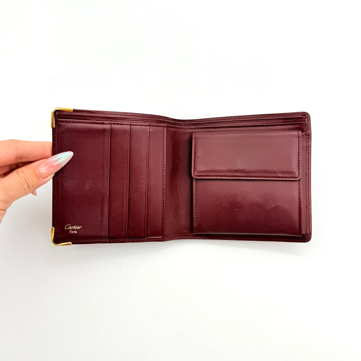 Cartier Must de Cartier bifold wallet in burgundy leather with gold-tone corners and embossed logo, photographed against a white background.