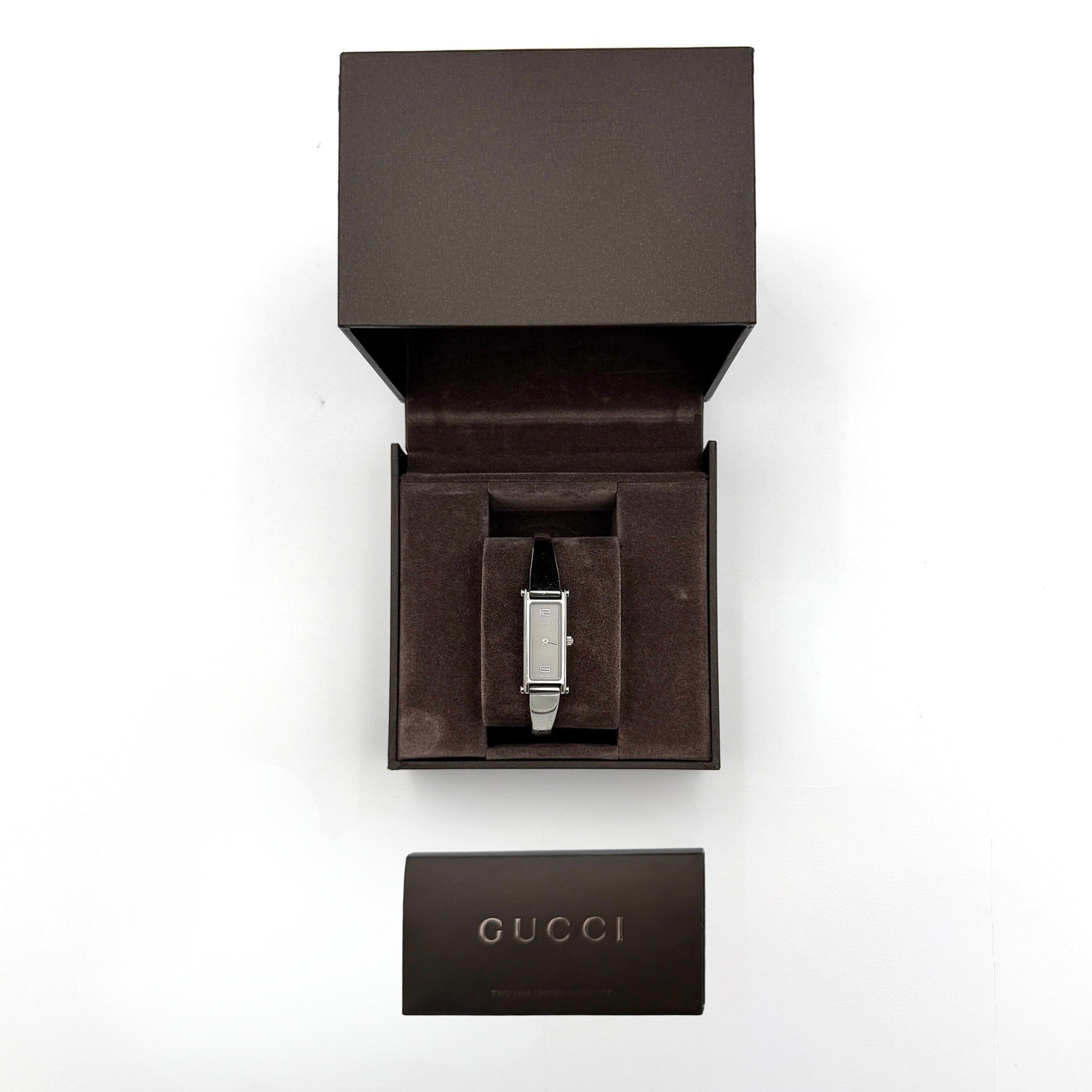The Gucci 1500L Grey Dial Watch rests elegantly on its plush brown cushion atop a branded box, showcasing its sleek rectangular design and polished silver-tone bracelet.