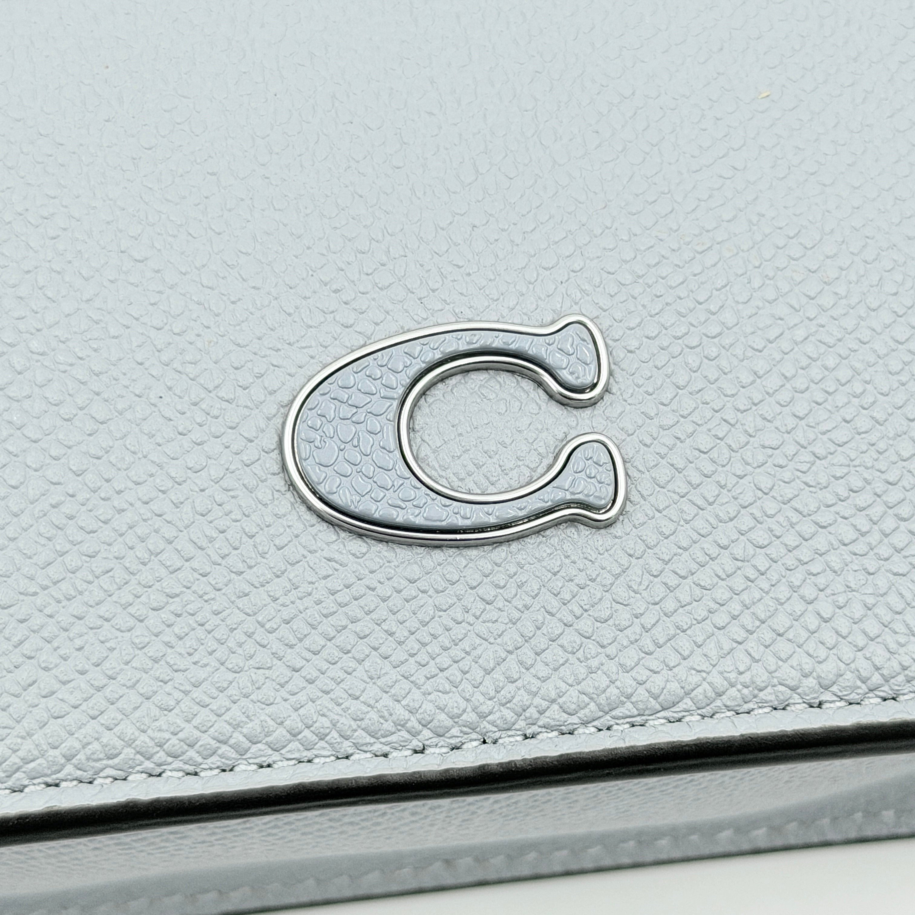 Sky blue Coach grained leather shoulder bag with silver-tone C plaque and short strap, displayed against a white background.