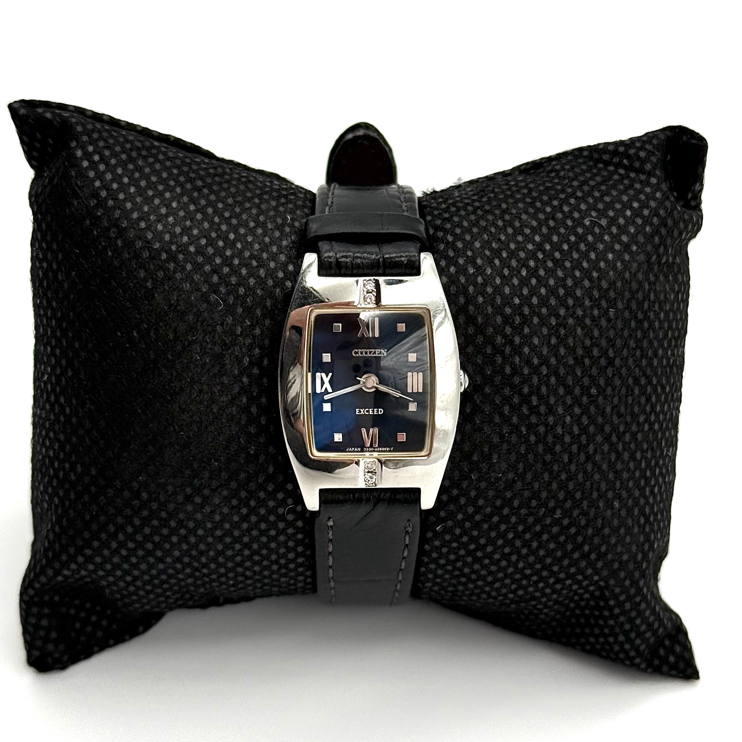 Citizen Exceed watch featuring a dark blue dial, tonneau-shaped stainless steel case, and black leather strap displayed on a black cushion.