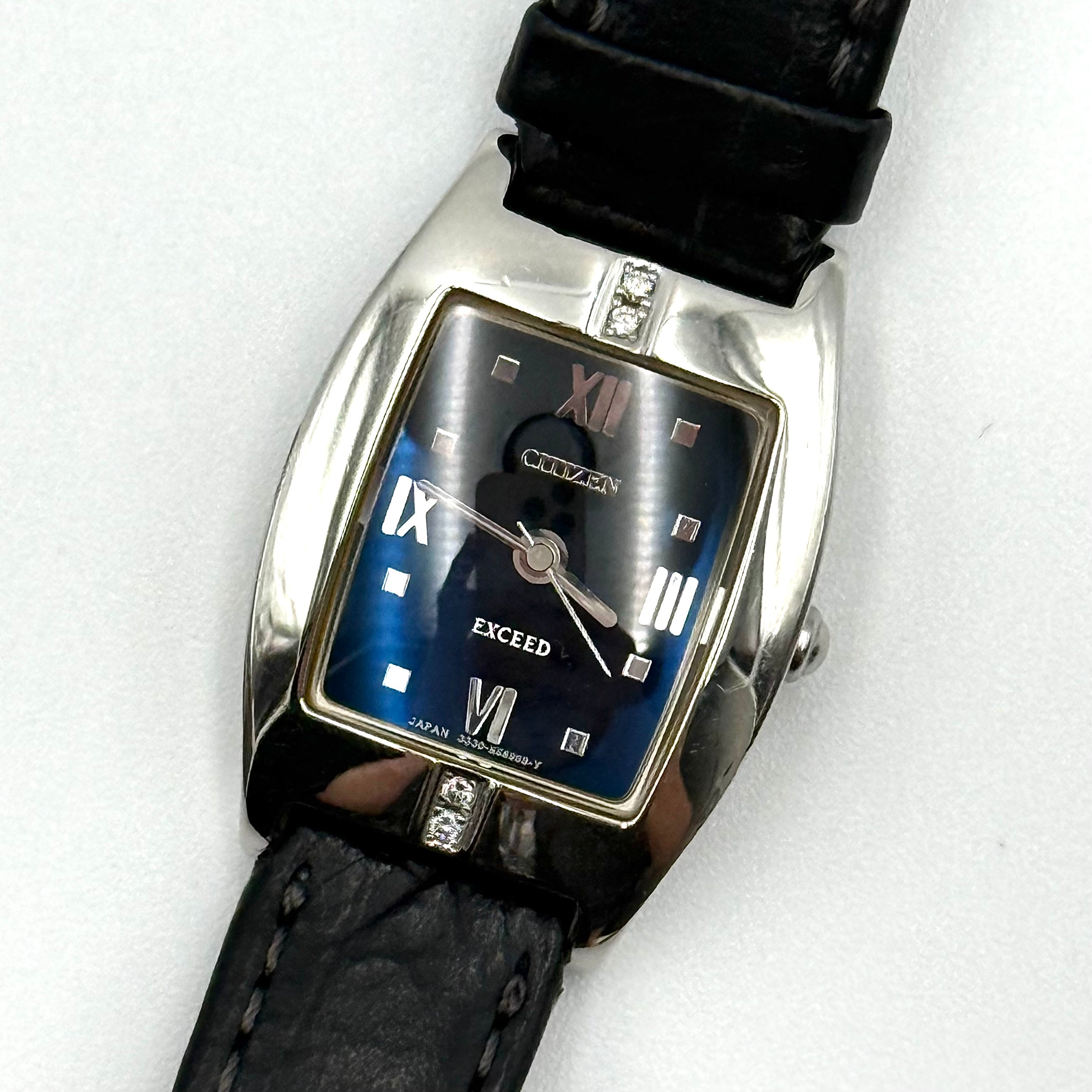 Citizen Exceed watch featuring a dark blue dial, tonneau-shaped stainless steel case, and black leather strap displayed on a black cushion.