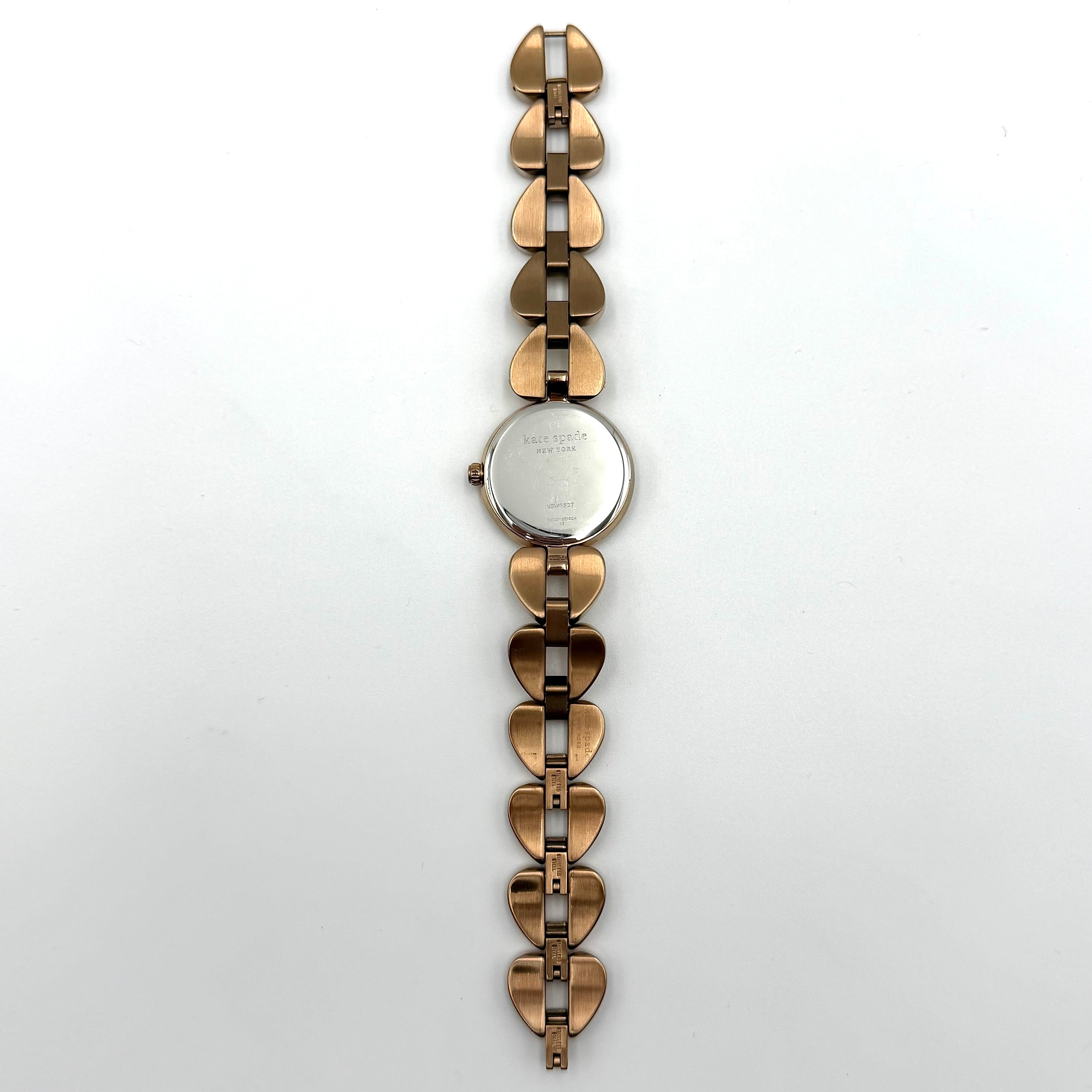 Kate Spade Annadale gold bracelet watch with white dial and heart-shaped links displayed on a black cushion.