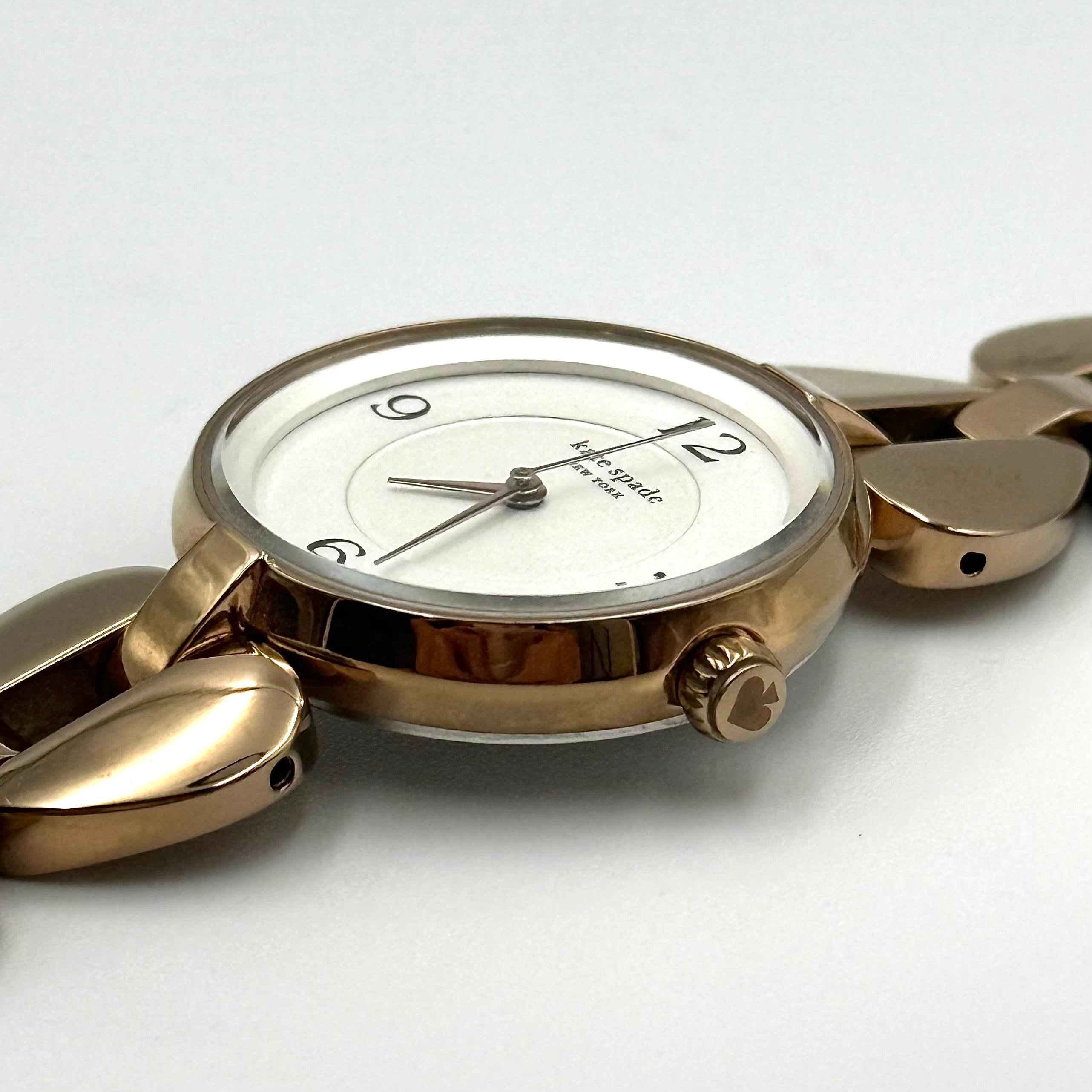 Kate Spade Annadale gold bracelet watch with white dial and heart-shaped links displayed on a black cushion.