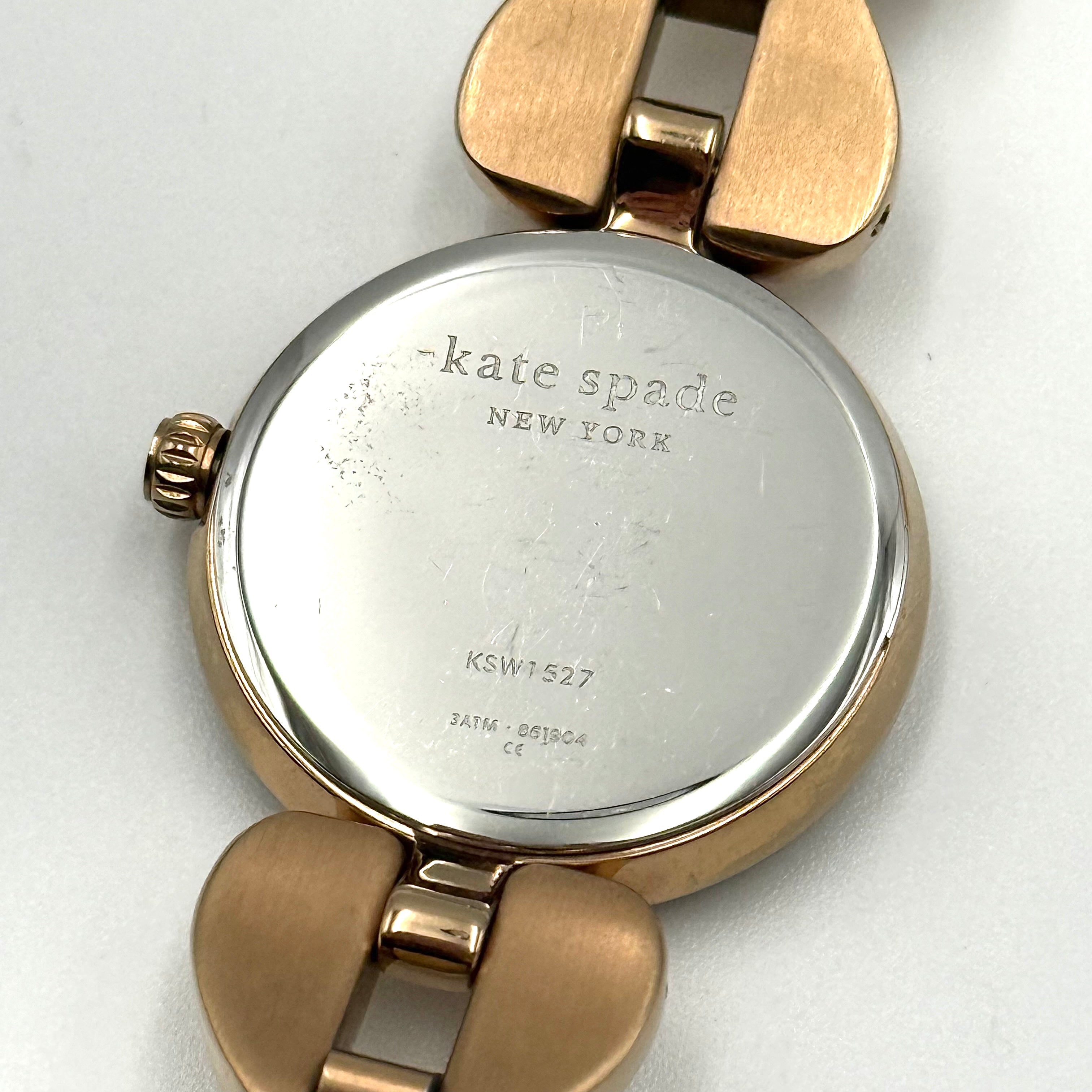 Kate Spade Annadale gold bracelet watch with white dial and heart-shaped links displayed on a black cushion.
