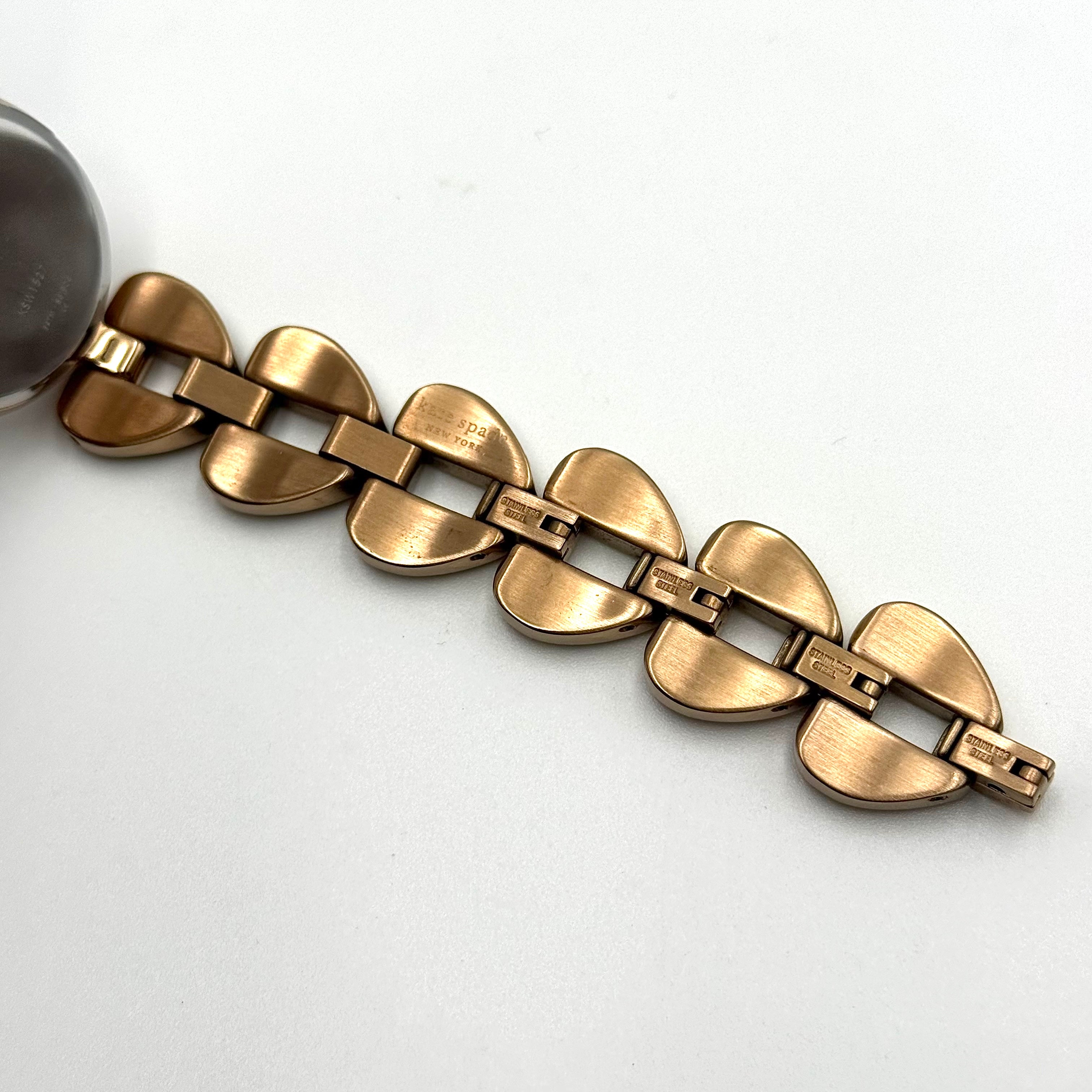 Kate Spade Annadale gold bracelet watch with white dial and heart-shaped links displayed on a black cushion.