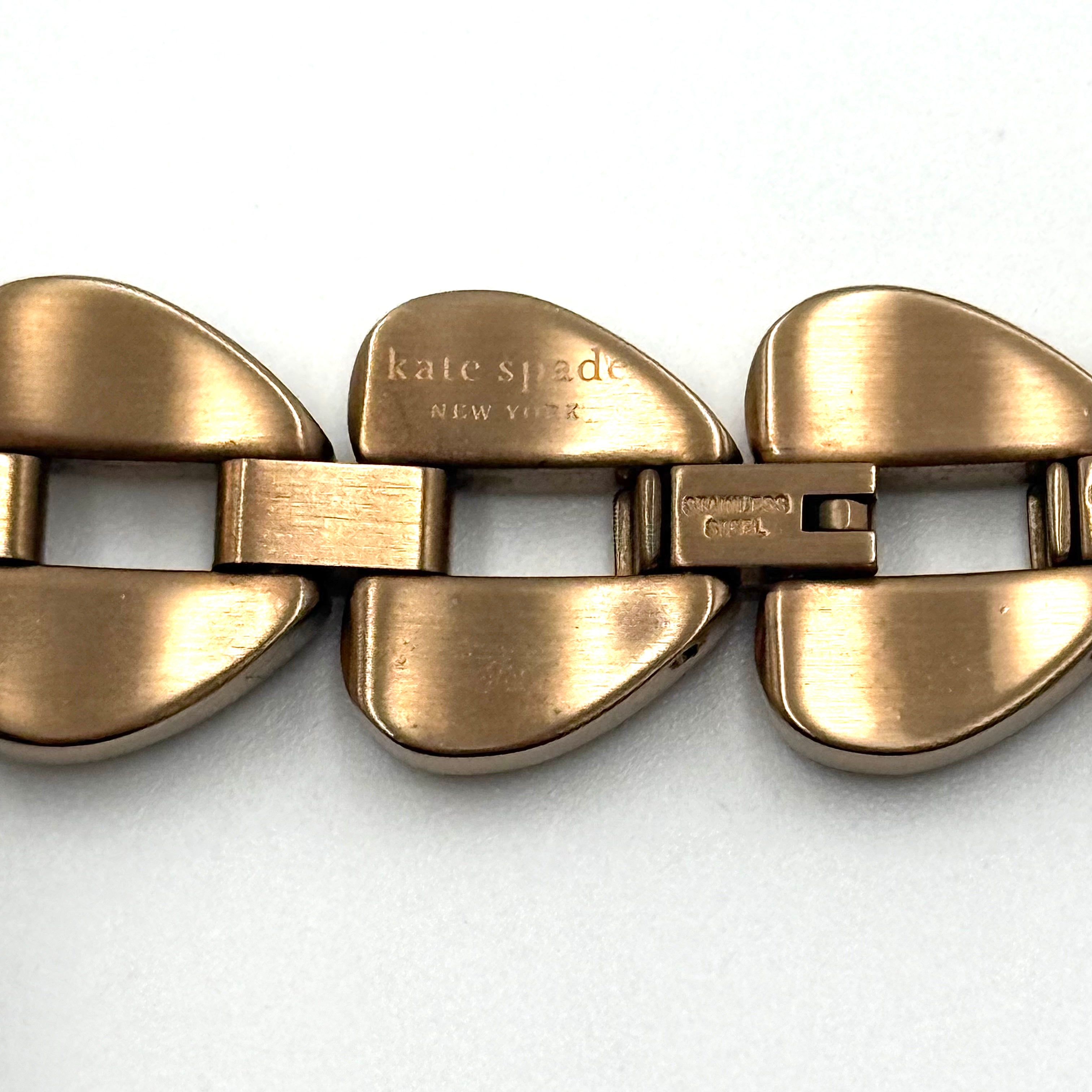 Kate Spade Annadale gold bracelet watch with white dial and heart-shaped links displayed on a black cushion.