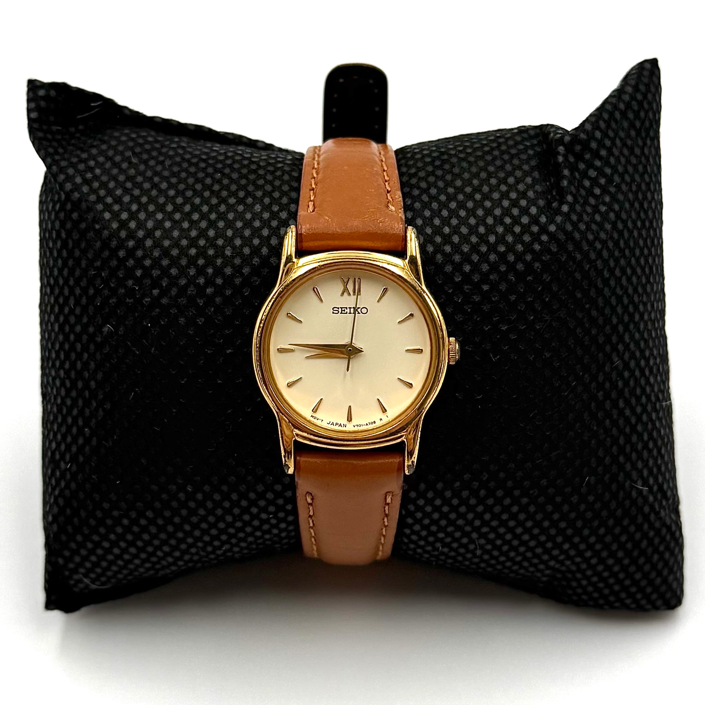 Seiko V701-A110 quartz watch with gold-tone case, cream dial, and brown leather strap on a display pillow.