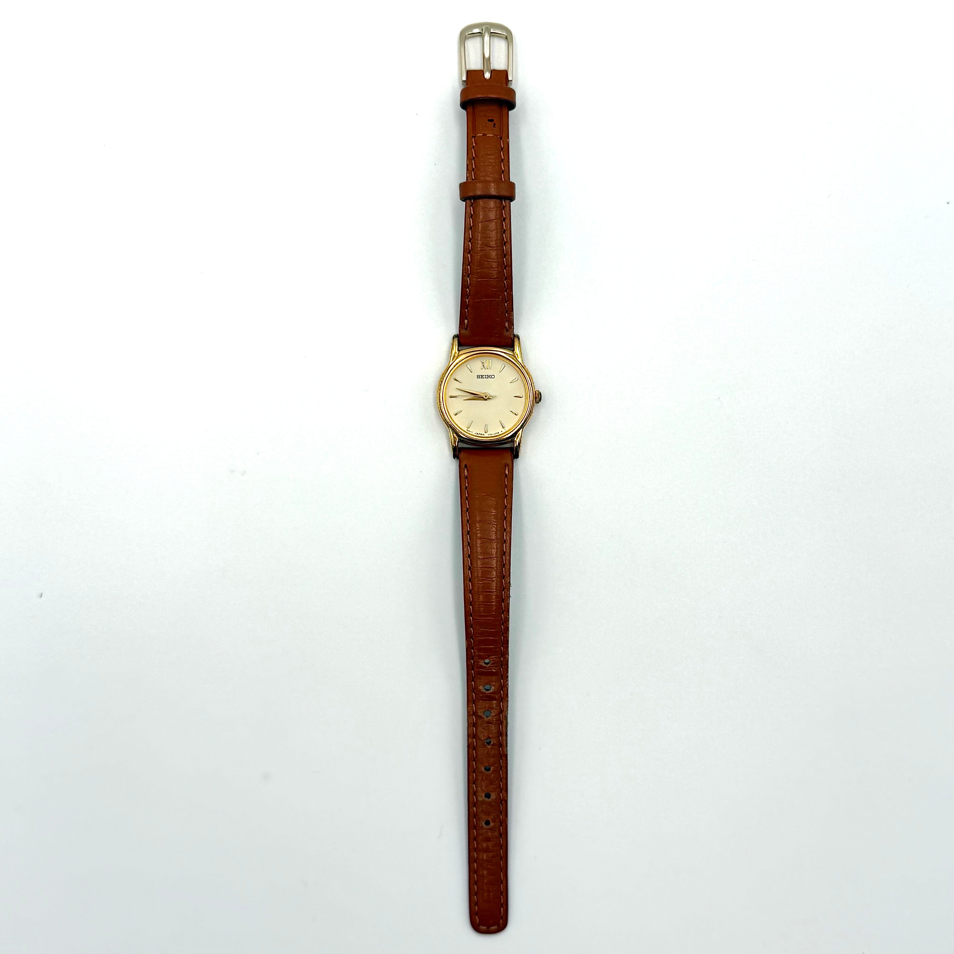 Seiko V701-A110 quartz watch with gold-tone case, cream dial, and brown leather strap on a display pillow.