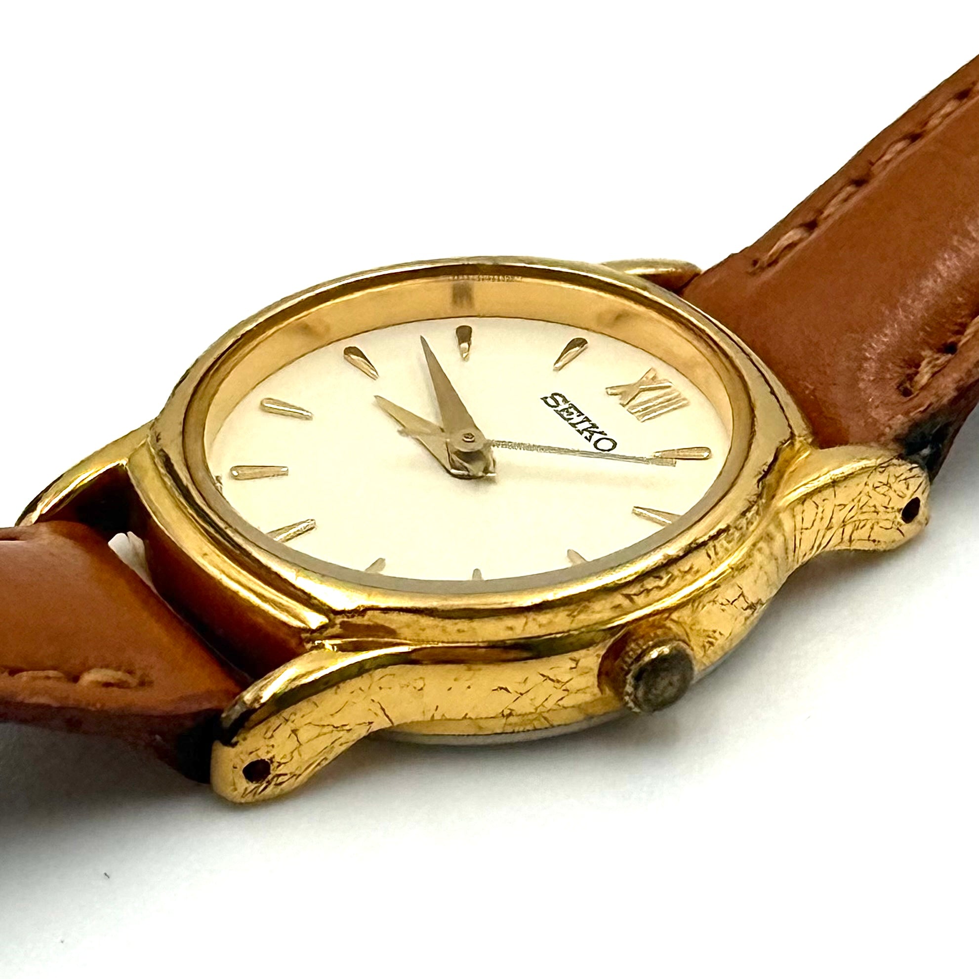 Seiko V701-A110 quartz watch with gold-tone case, cream dial, and brown leather strap on a display pillow.