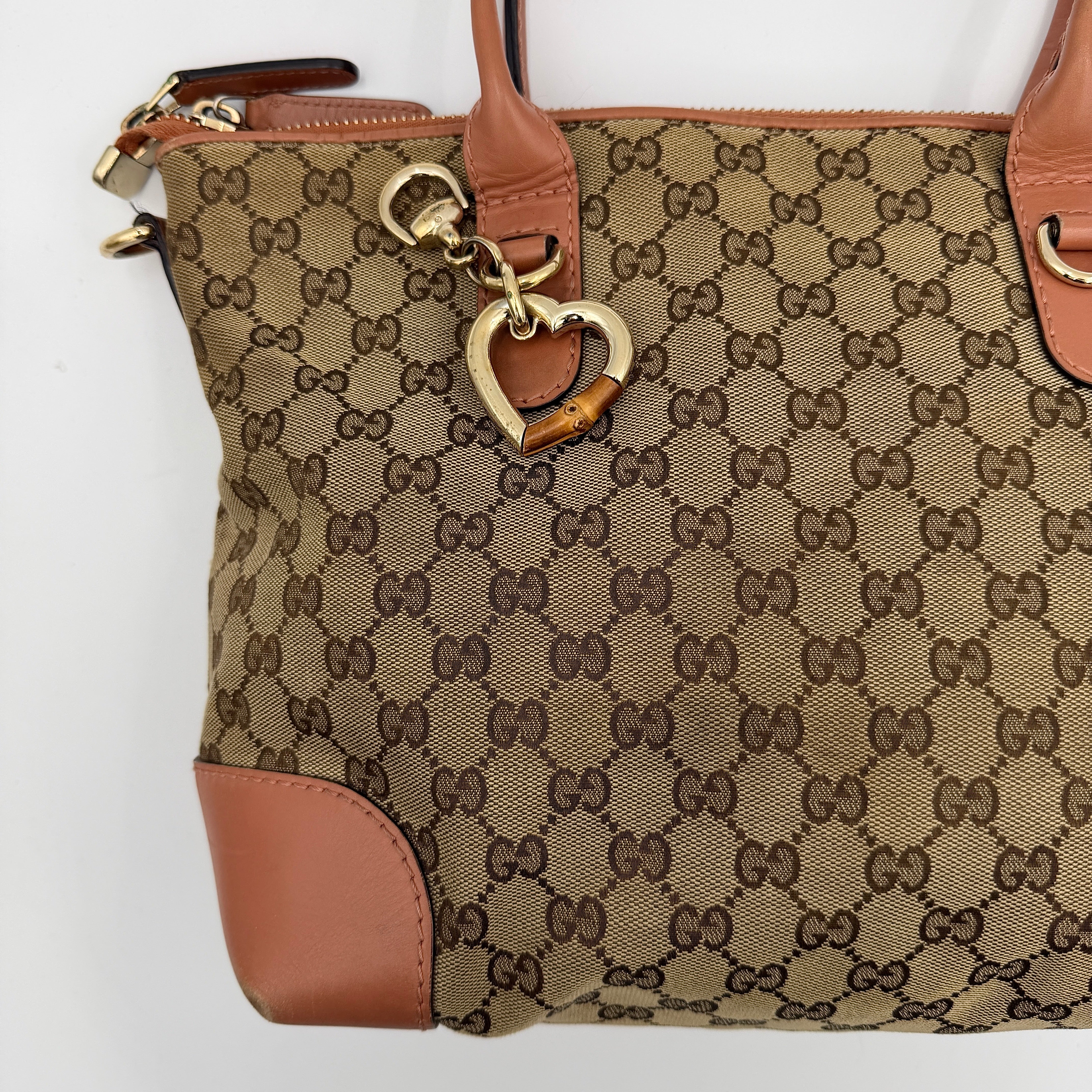 Gucci beige GG monogram shoulder bag with orange leather trim, heart bit charm, and detachable strap displayed on a white background.