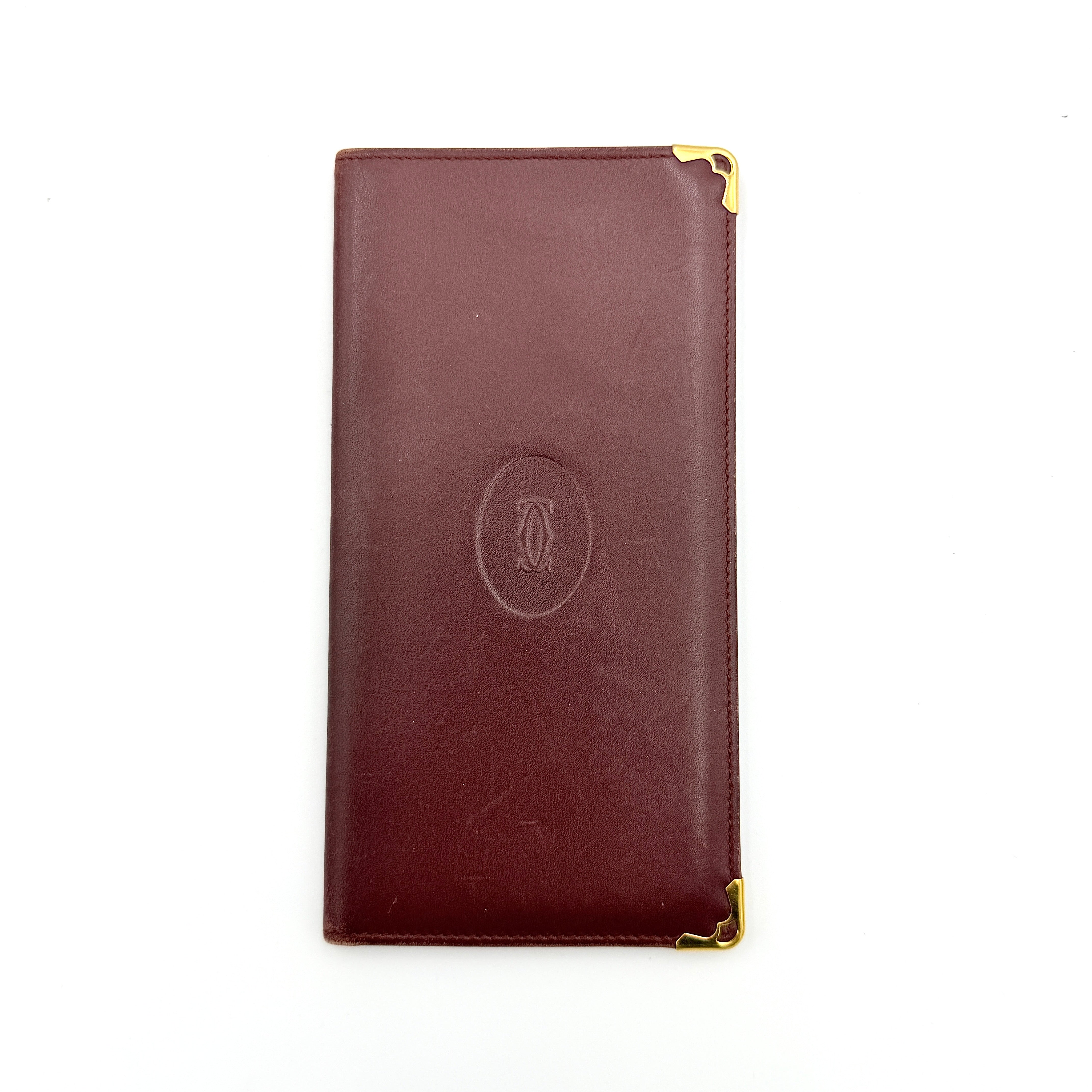 The Cartier Must de Cartier long bifold wallet in rich bordeaux calfskin with gold-tone corner accents and an embossed logo.
