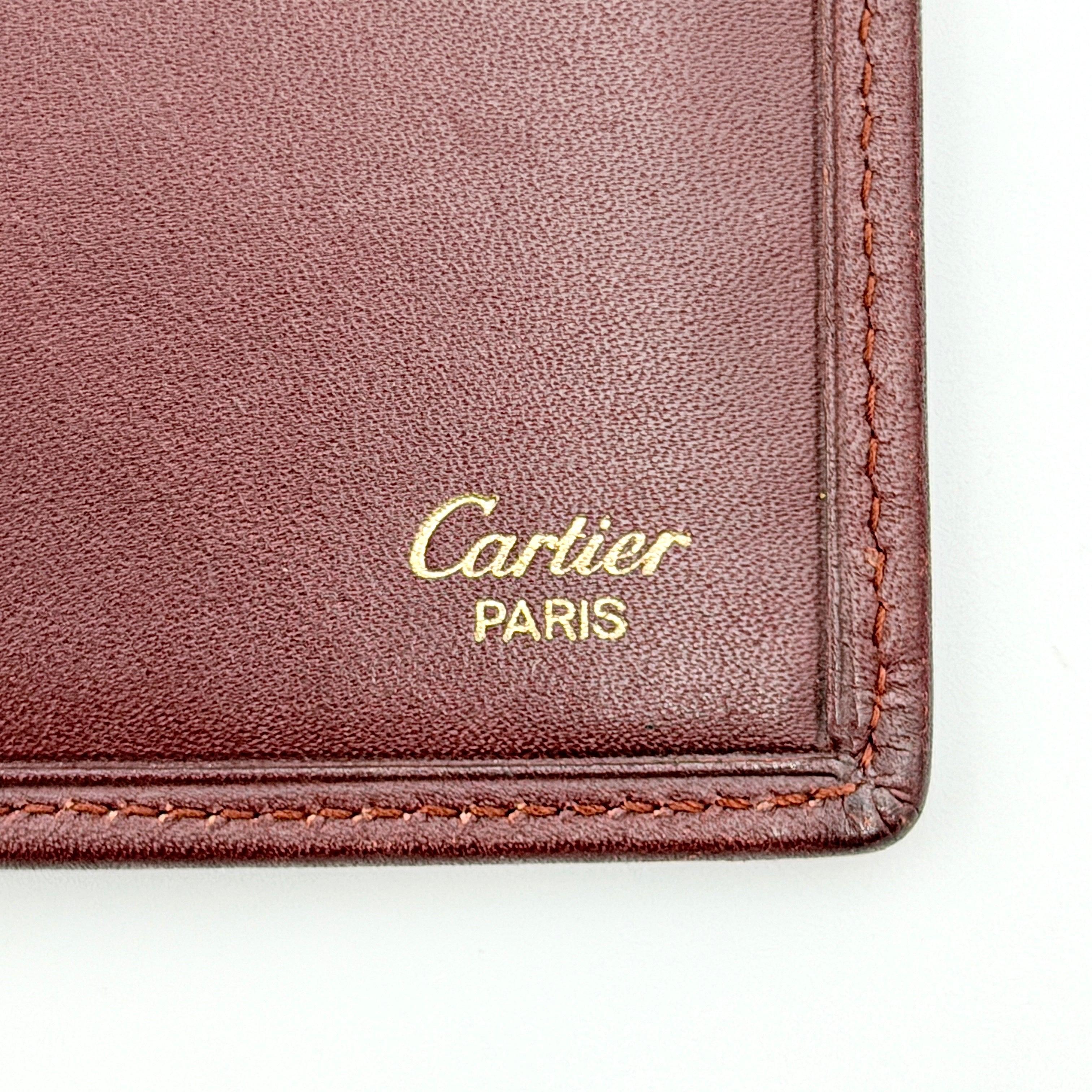 The Cartier Must de Cartier long bifold wallet in rich bordeaux calfskin with gold-tone corner accents and an embossed logo.