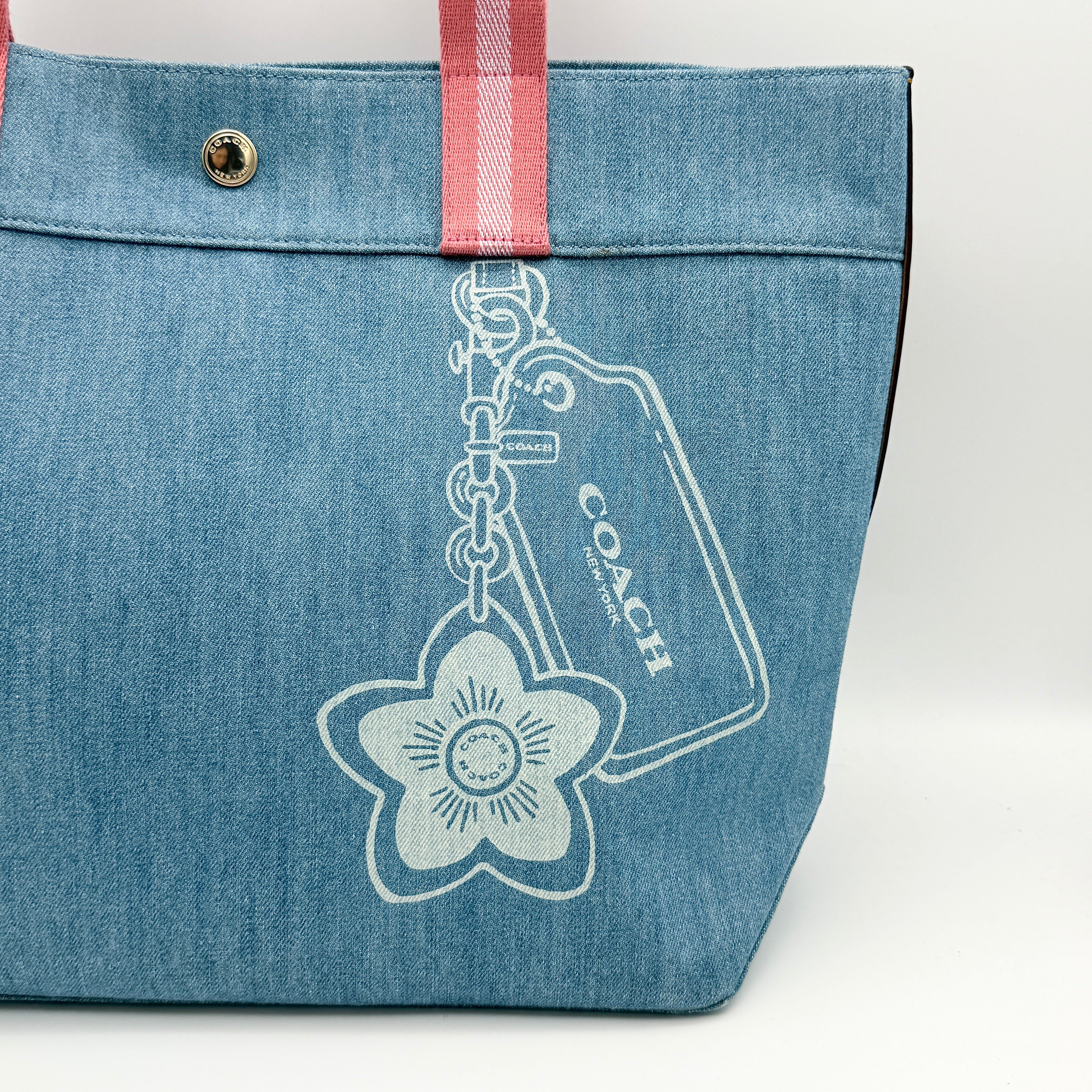 Coach denim tote bag with pink handles and a white flower and hang tag graphic at the corner