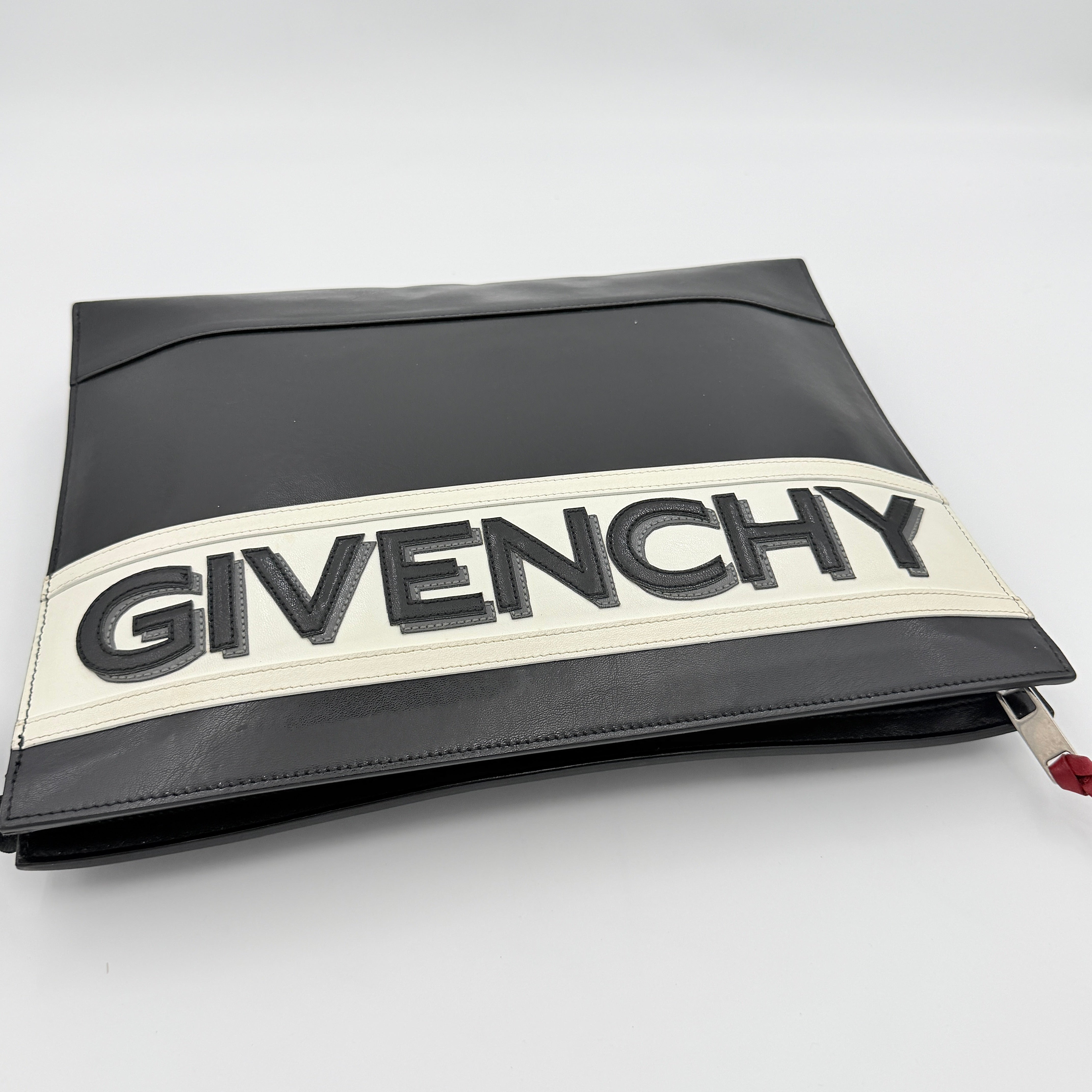 Front view of a Givenchy black leather clutch with bold white logo panel and red zip pull.