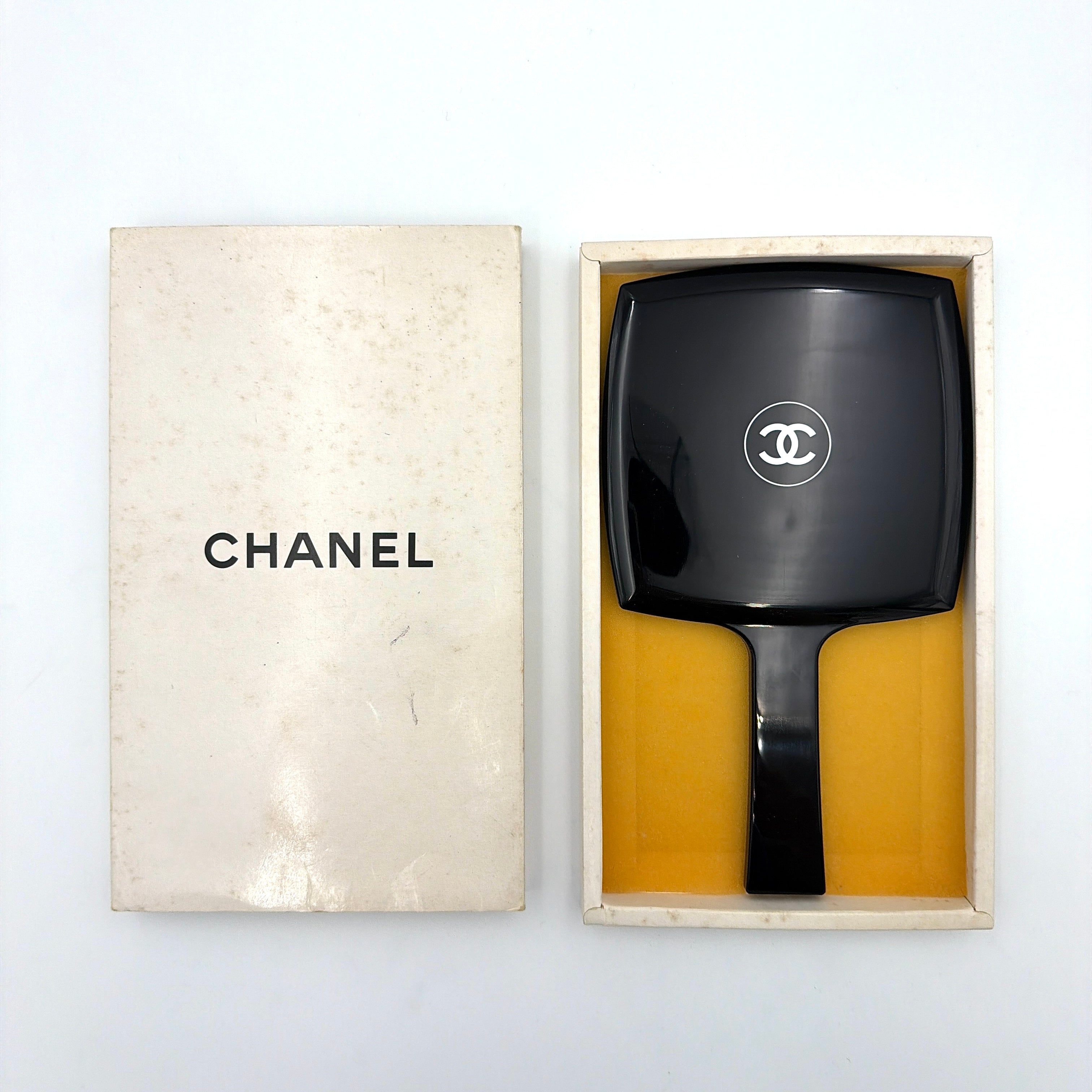 Chanel black hand mirror with white double C logo, presented in its original box with a yellow interior lining.