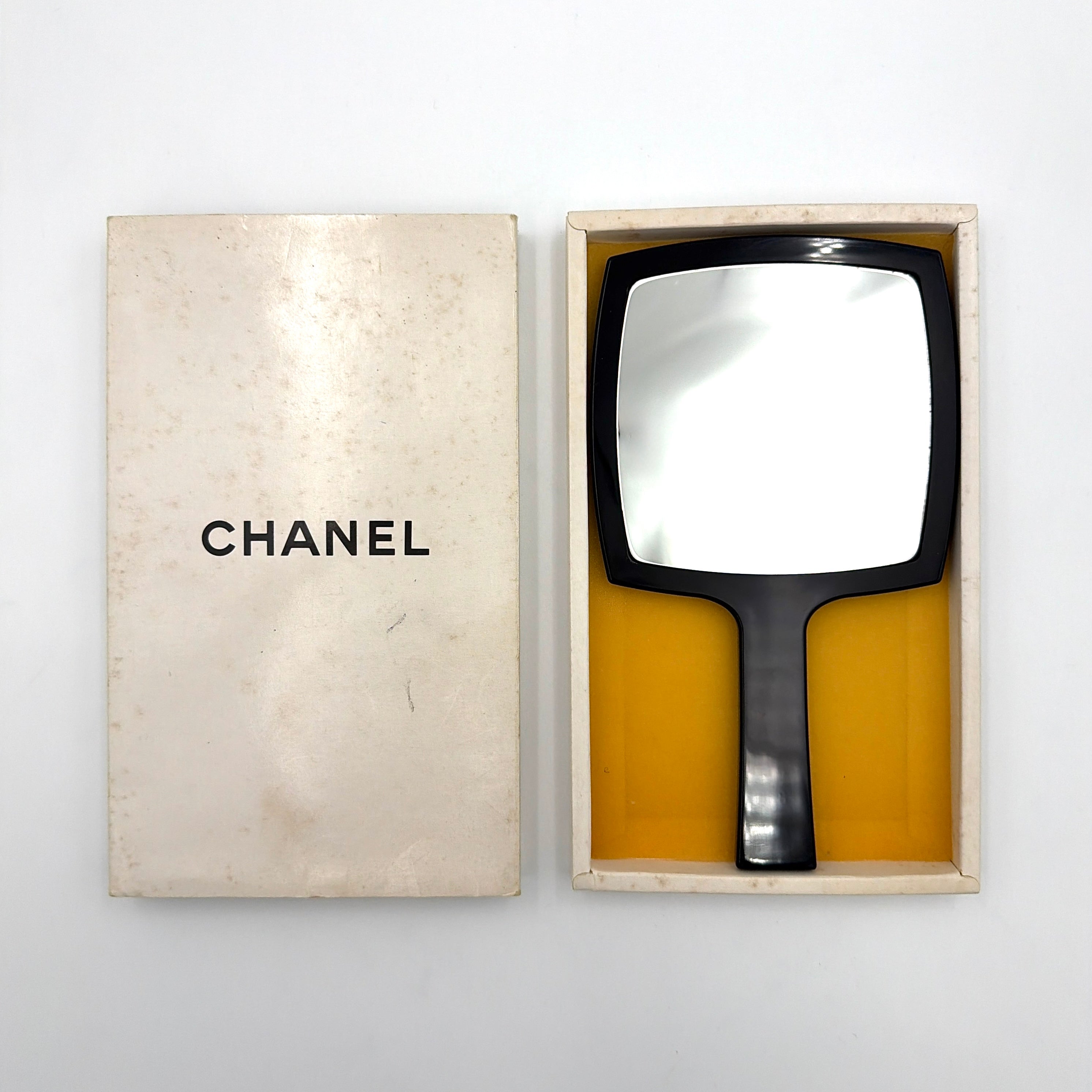 Chanel black hand mirror with white double C logo, presented in its original box with a yellow interior lining.