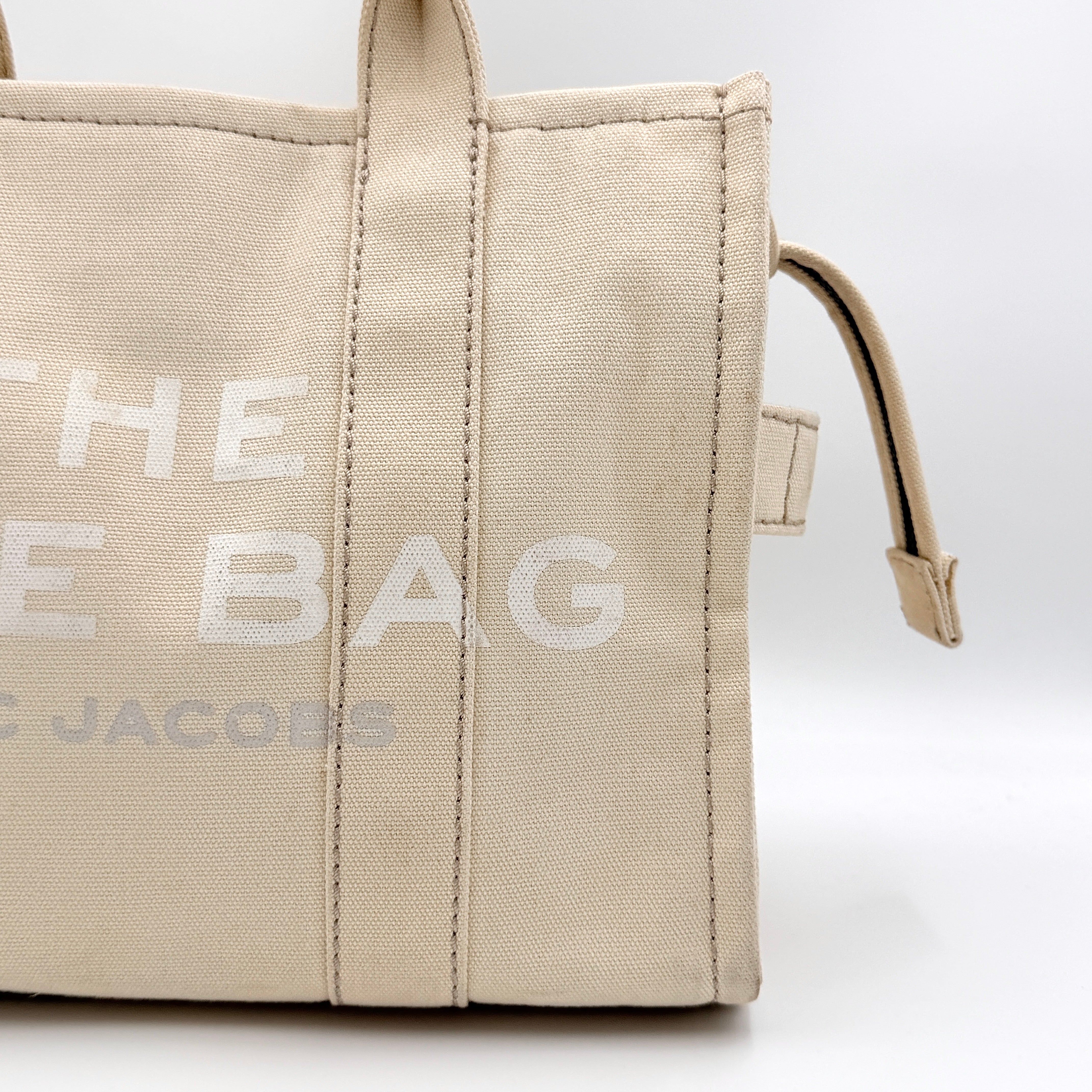 Marc Jacobs The Canvas Tote Bag in beige canvas with bold text, featuring top handles and zip closure.