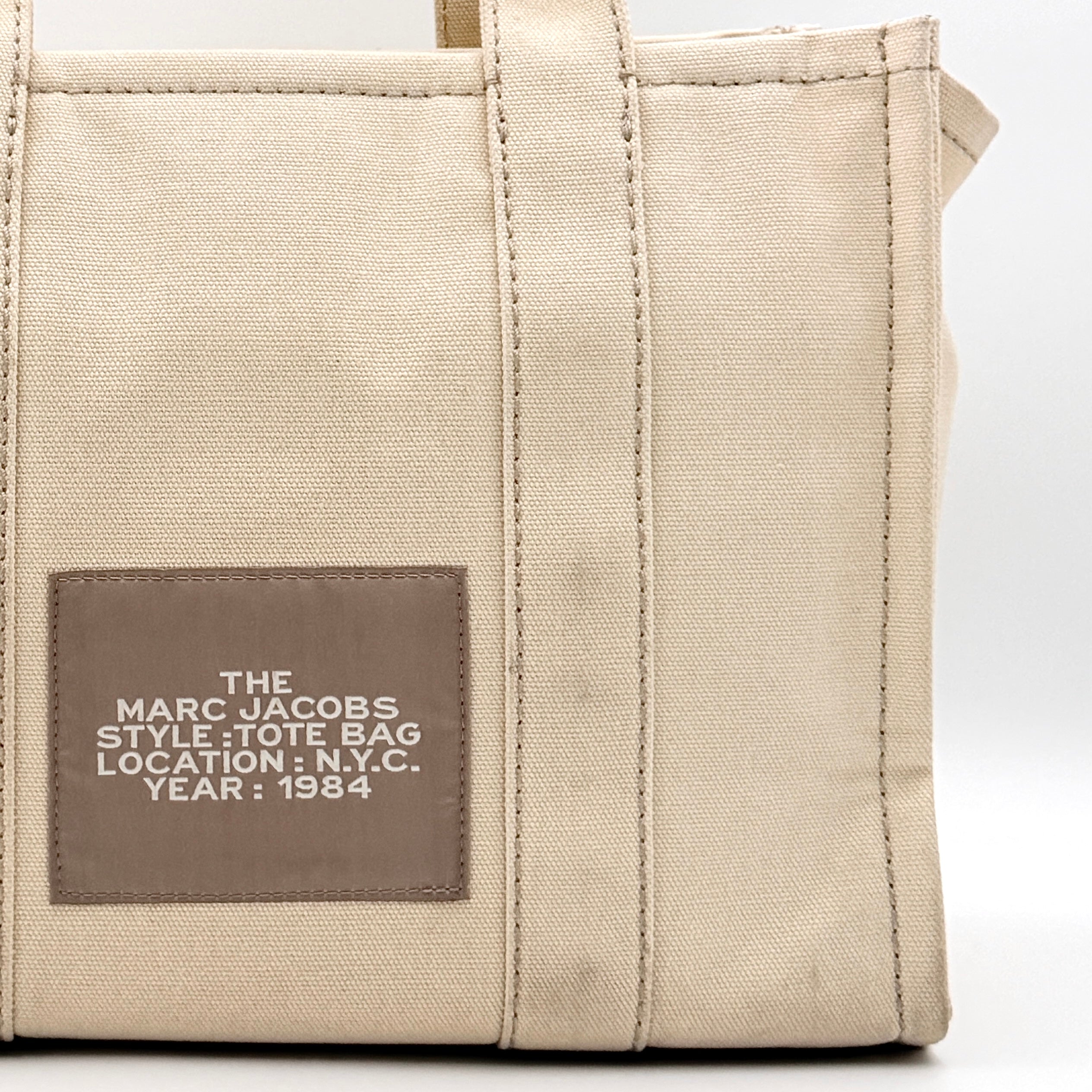 Marc Jacobs The Canvas Tote Bag in beige canvas with bold text, featuring top handles and zip closure.