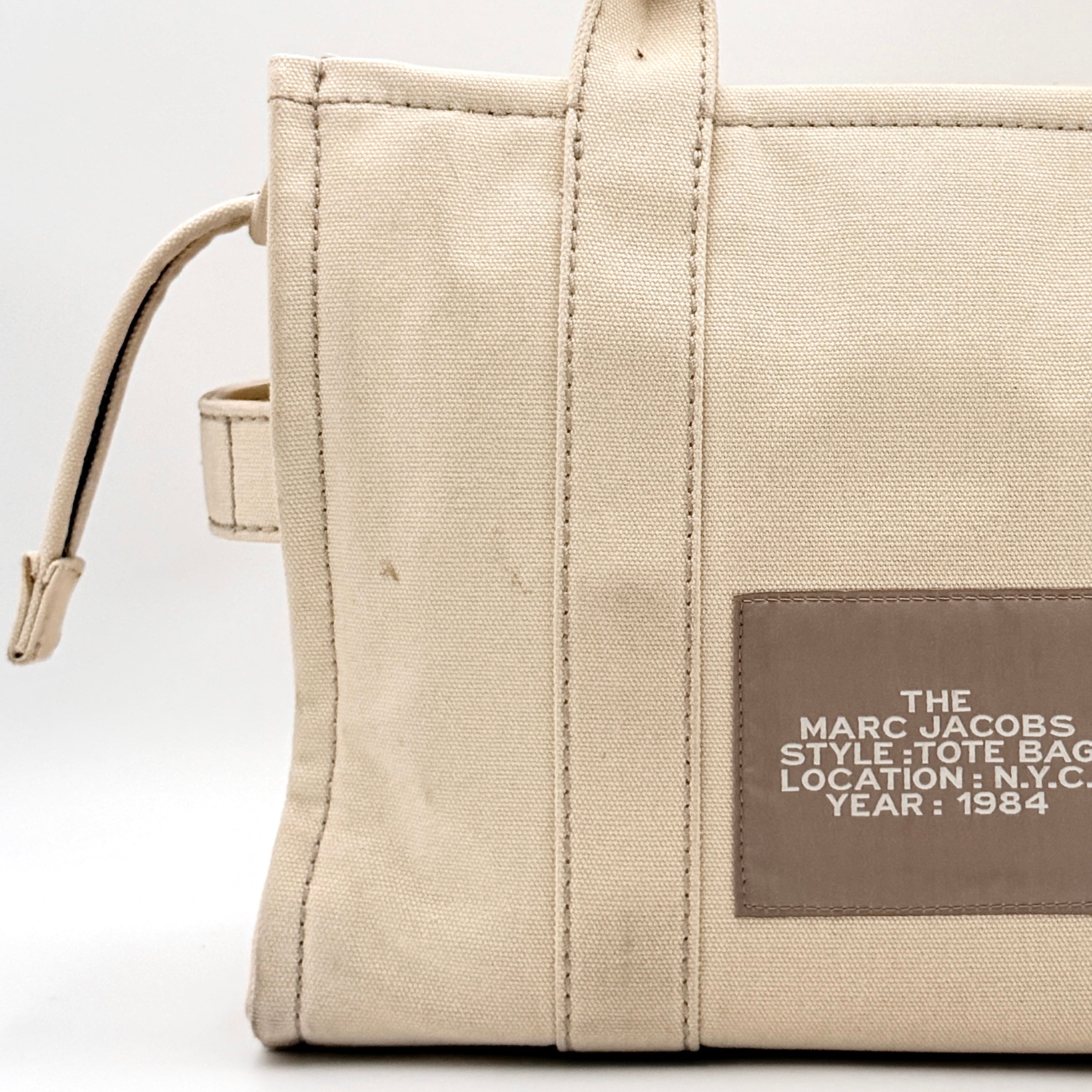 Marc Jacobs The Canvas Tote Bag in beige canvas with bold text, featuring top handles and zip closure.