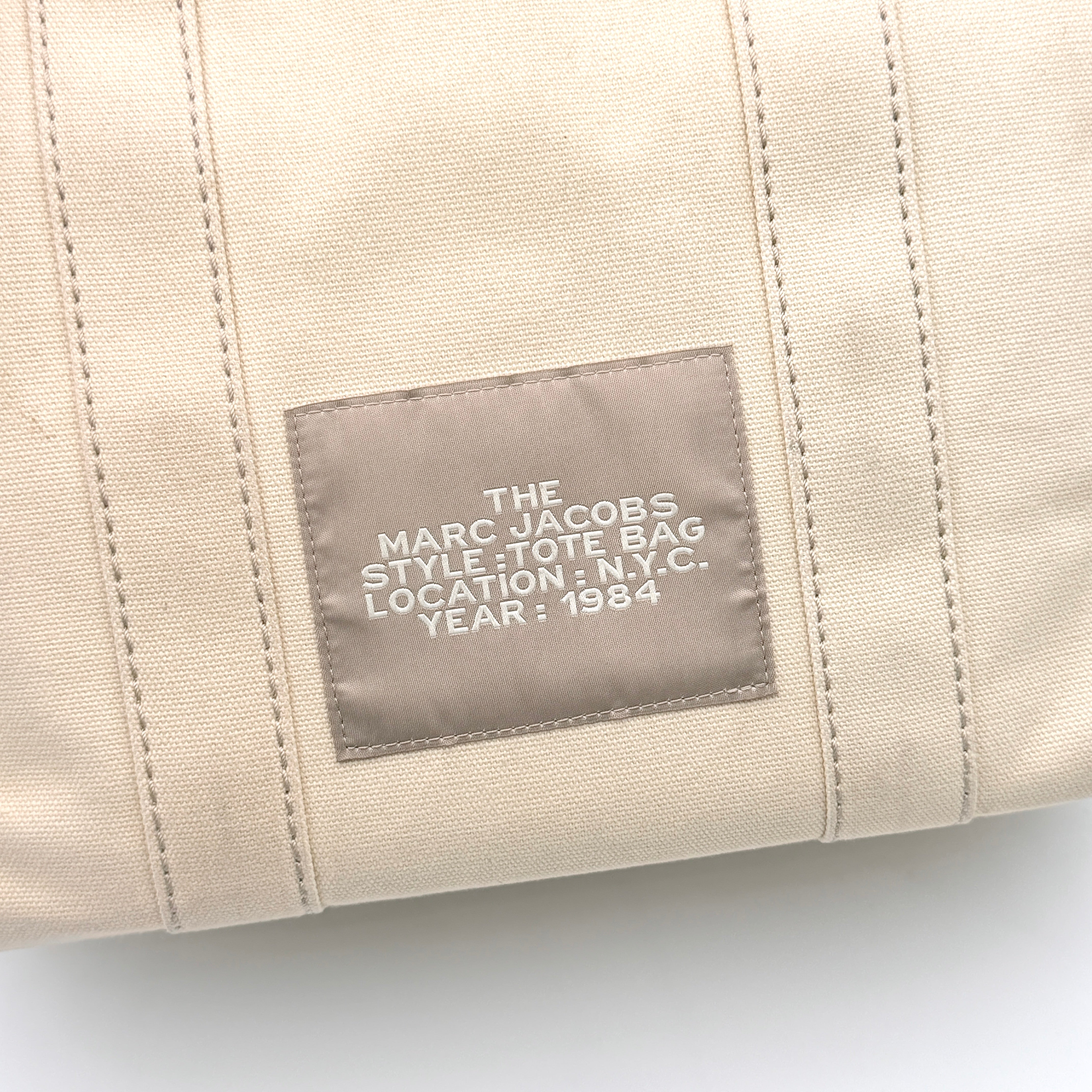 Marc Jacobs The Canvas Tote Bag in beige canvas with bold text, featuring top handles and zip closure.