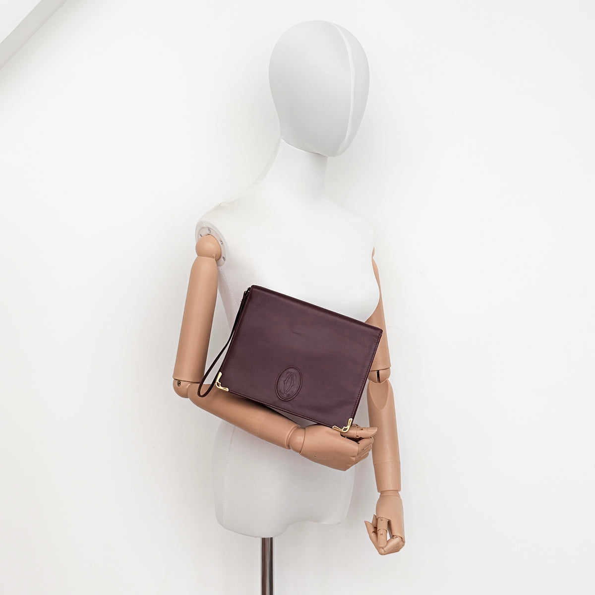 Cartier Must de Cartier wristlet clutch in burgundy leather with gold-tone corners and wrist strap, pictured with authenticity card and booklet.