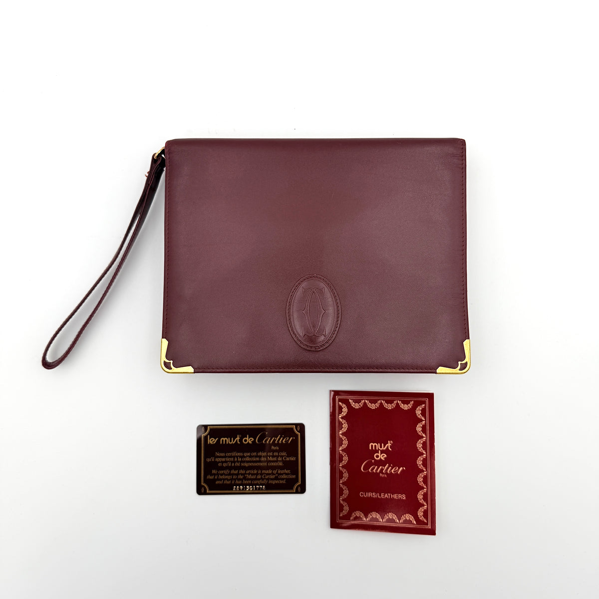 Cartier Must de Cartier wristlet clutch in burgundy leather with gold-tone corners and wrist strap, pictured with authenticity card and booklet.