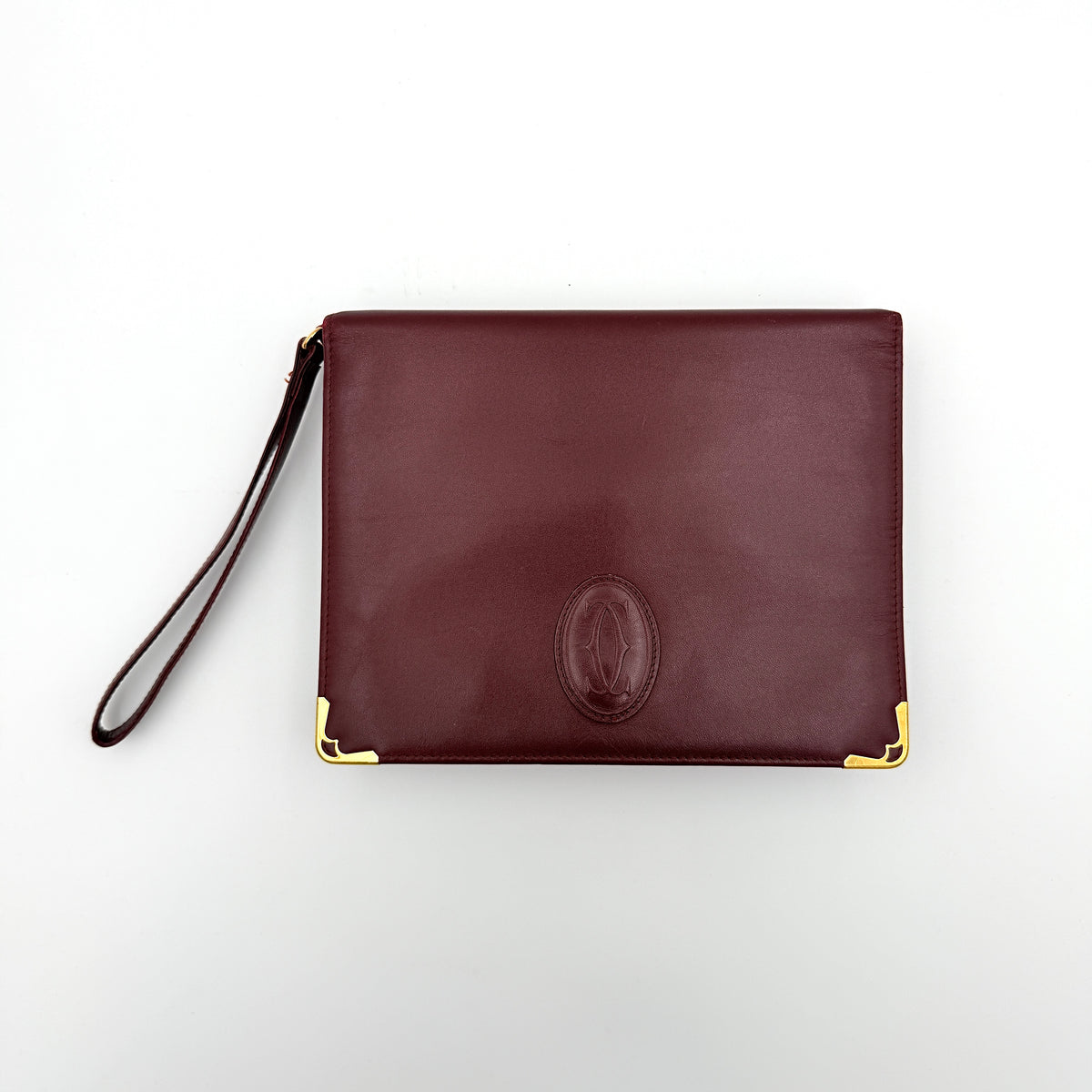 Cartier Must de Cartier wristlet clutch in burgundy leather with gold-tone corners and wrist strap, pictured with authenticity card and booklet.