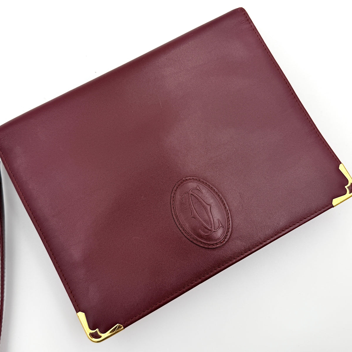 Cartier Must de Cartier wristlet clutch in burgundy leather with gold-tone corners and wrist strap, pictured with authenticity card and booklet.