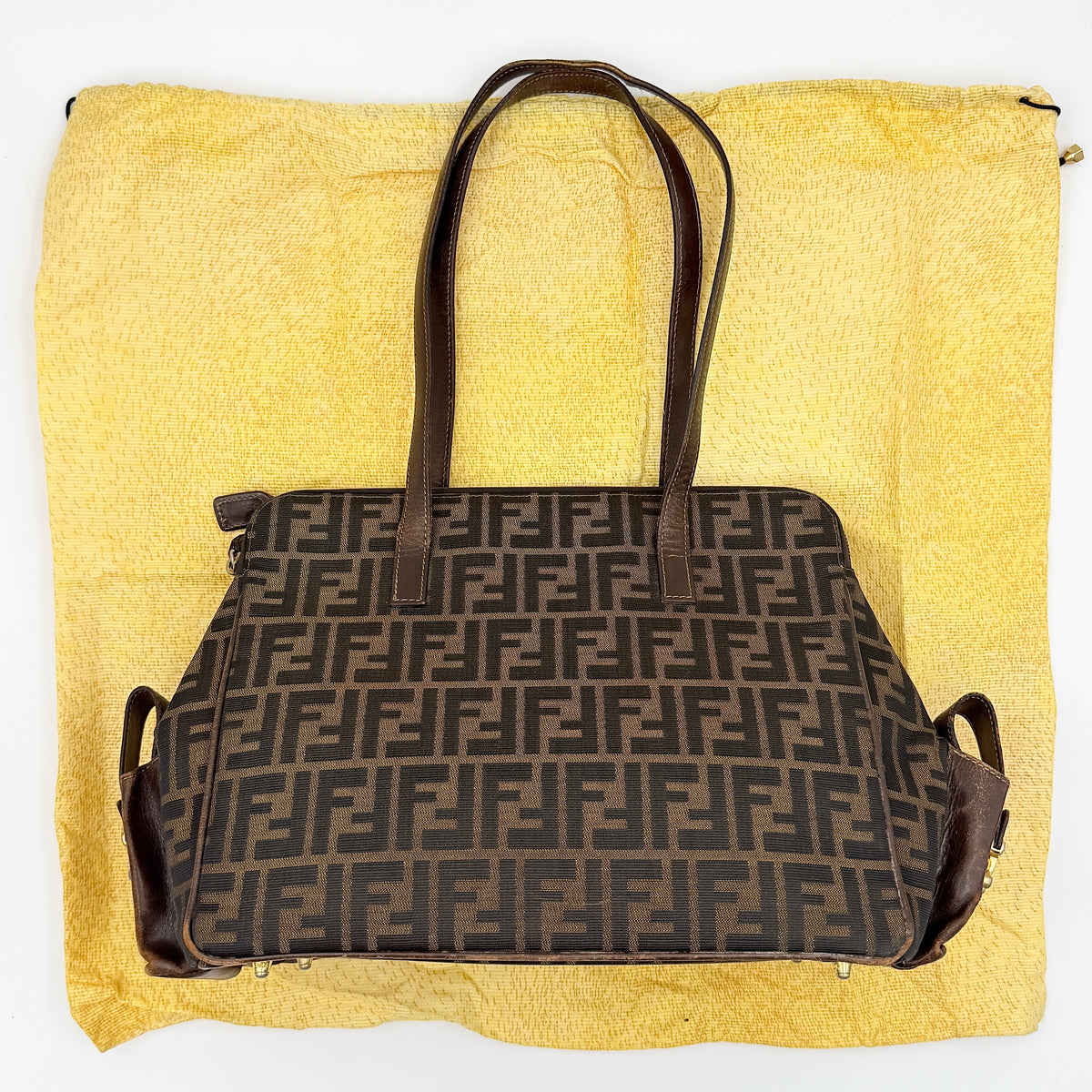 Fendi FF Zucca Double Zip Tote Bag in brown monogram canvas with leather straps and side pocket detail, shown on a yellow dust bag.