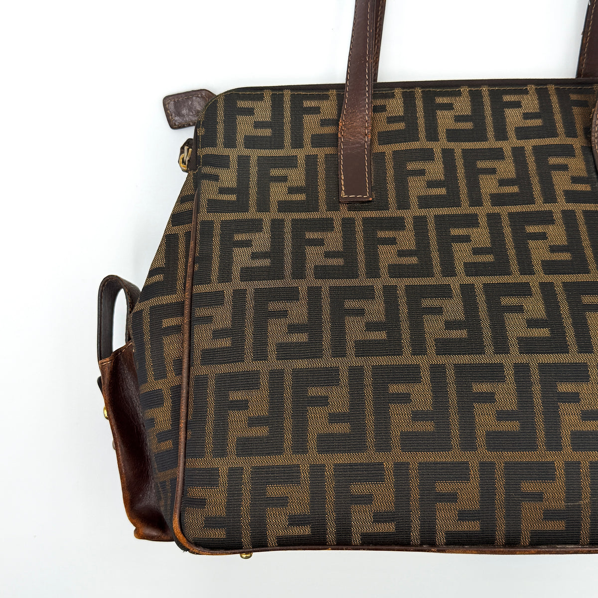 Fendi FF Zucca Double Zip Tote Bag in brown monogram canvas with leather straps and side pocket detail, shown on a yellow dust bag.