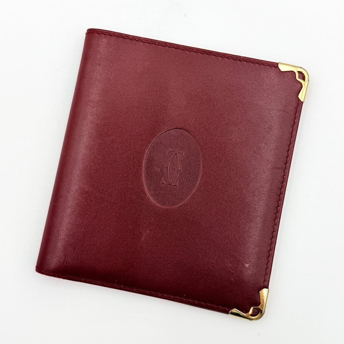 Cartier Must de Cartier bifold wallet in burgundy leather with gold-tone corner hardware and embossed logo, shown with authenticity card.