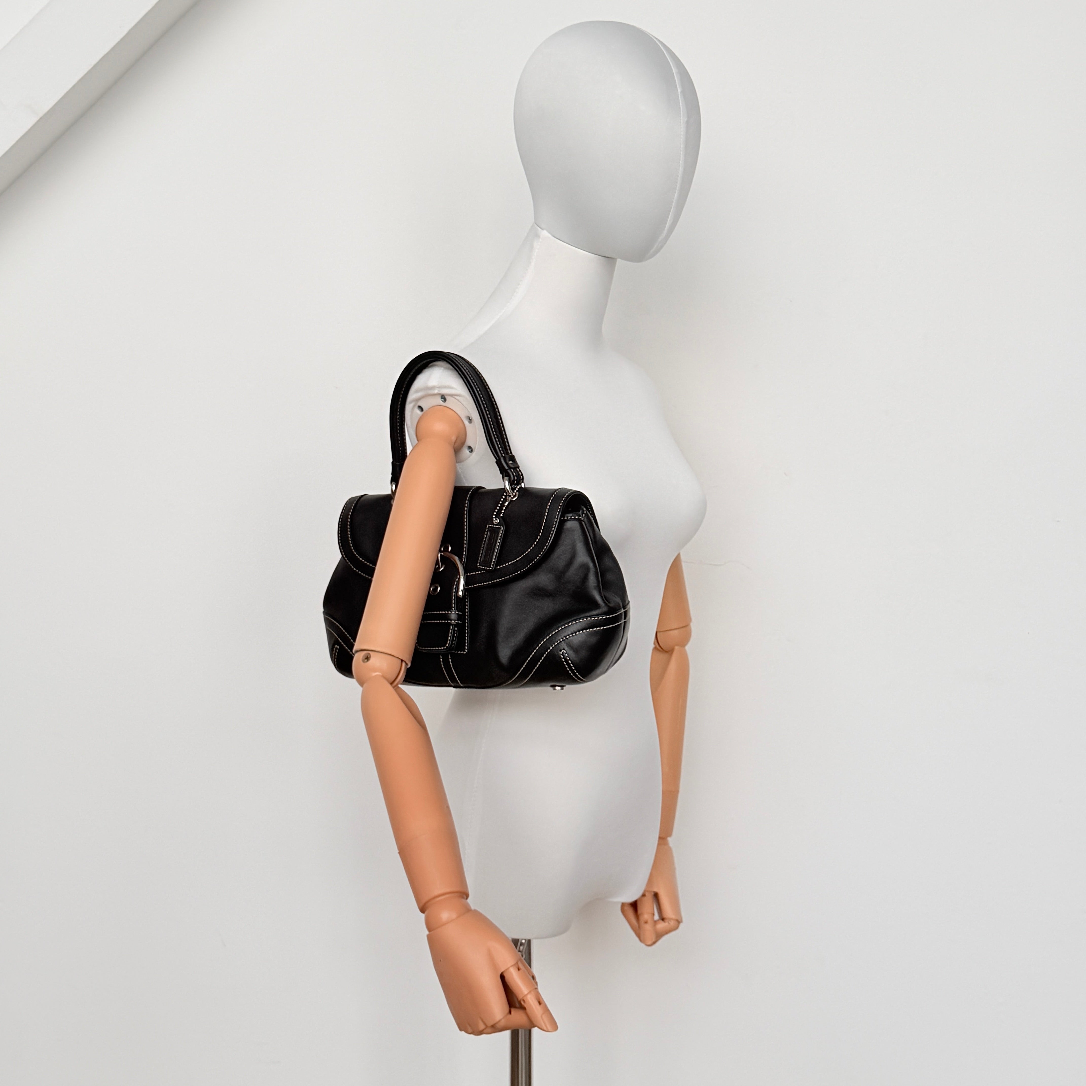 Coach Soho Buckle Top Handle Bag in black leather with contrast stitching and silver buckle, presented against a white background.