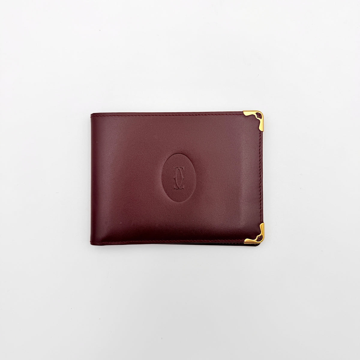 Cartier Must de Cartier burgundy leather bifold wallet with gold-tone corner accents shown against a white background.