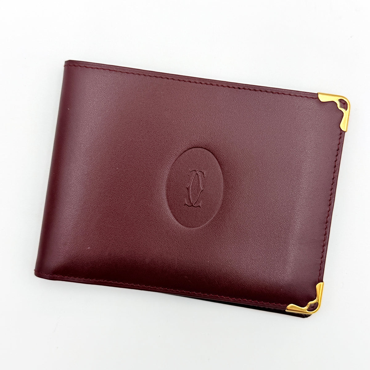Cartier Must de Cartier burgundy leather bifold wallet with gold-tone corner accents shown against a white background.