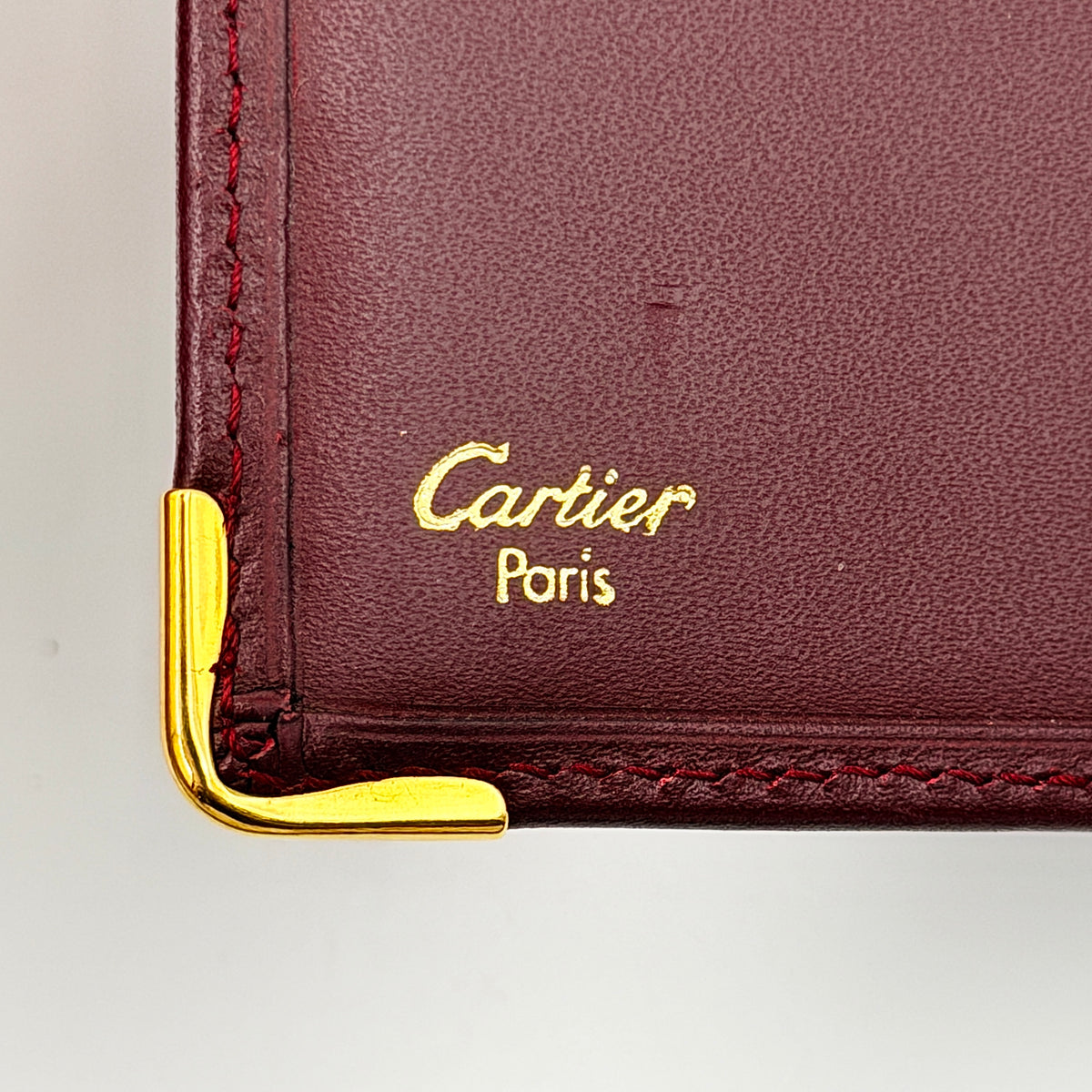 Cartier Must de Cartier burgundy leather bifold wallet with gold-tone corner accents shown against a white background.