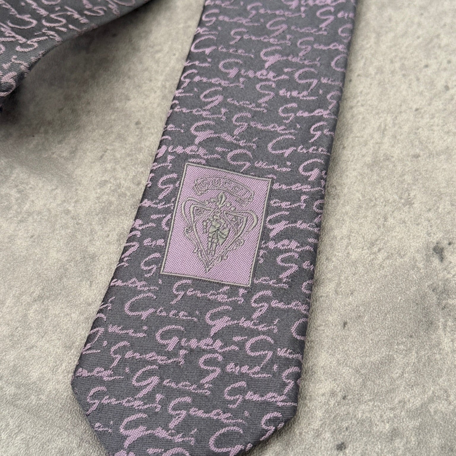 Vintage Gucci silk tie in gray with purple interlocking 'G' pattern and Gucci crest, exemplifying classic elegance and luxury style.