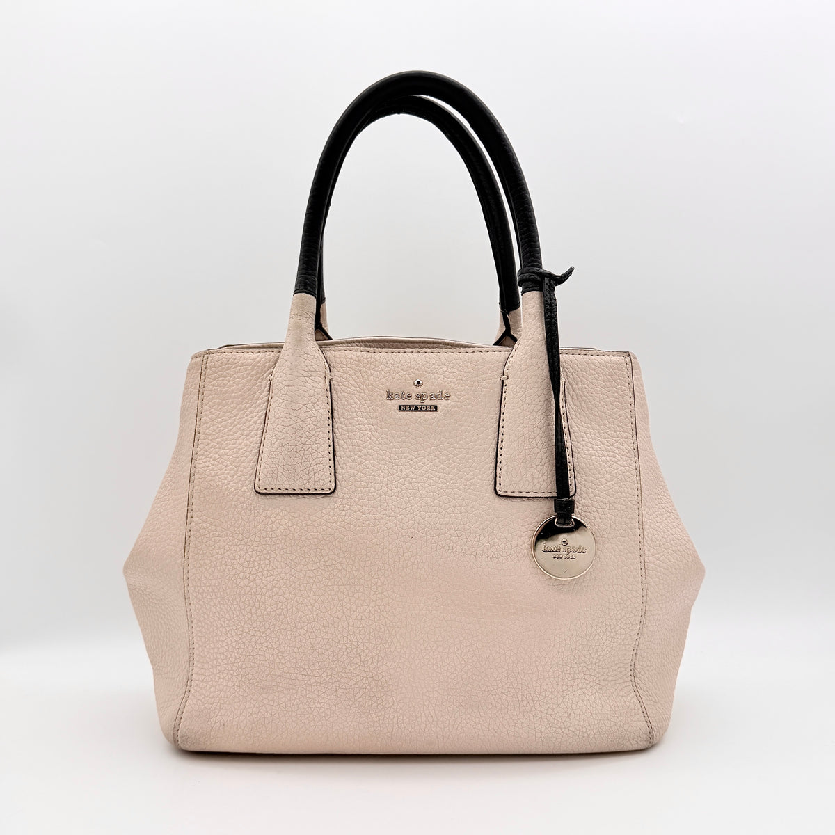 Kate Spade New York beige leather satchel with black handles, gold medallion, and grained texture against a white background.