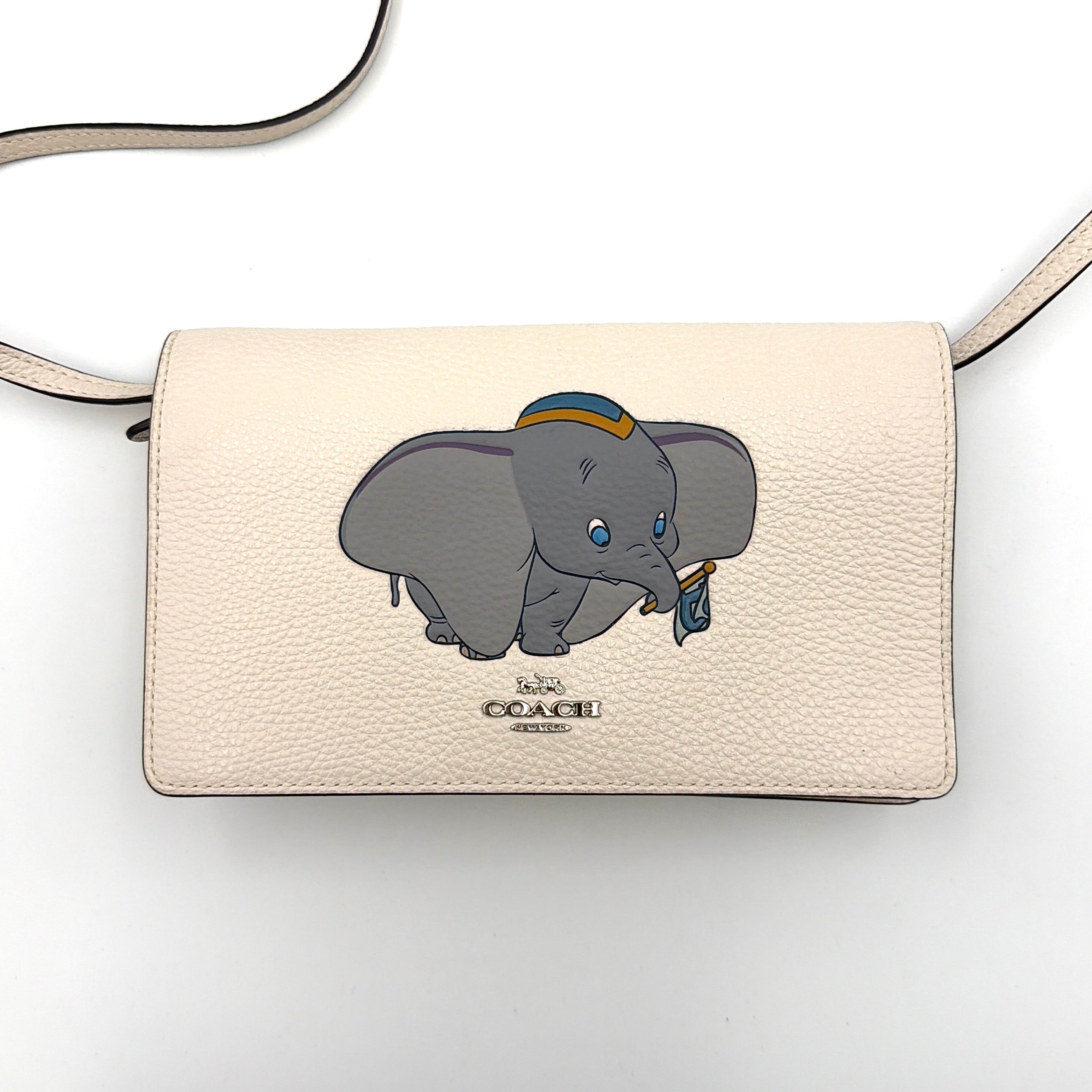 Coach x Disney Dumbo cream foldover crossbody bag with Dumbo illustration and slender strap, displayed against white background.