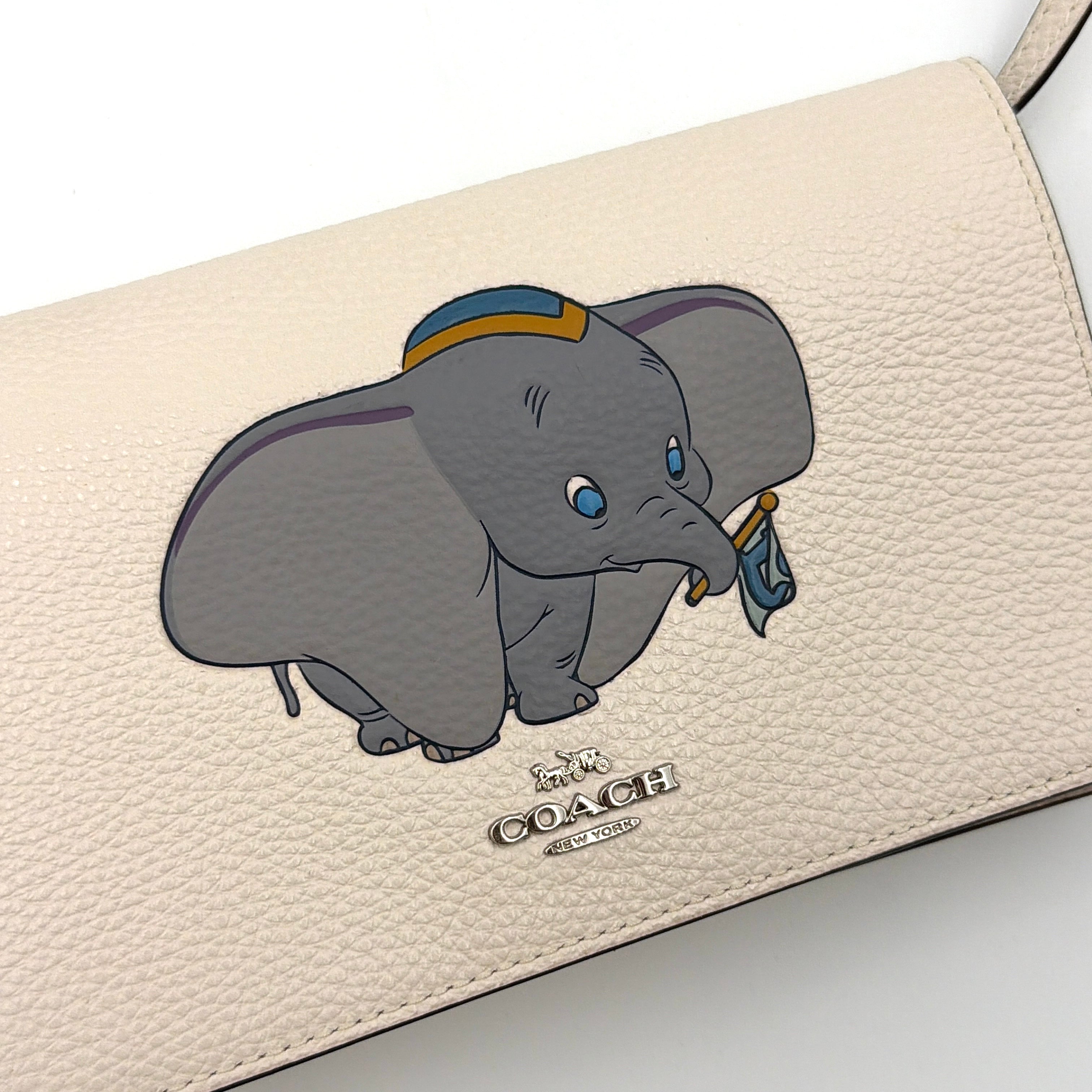 Coach x Disney Dumbo cream foldover crossbody bag with Dumbo illustration and slender strap, displayed against white background.
