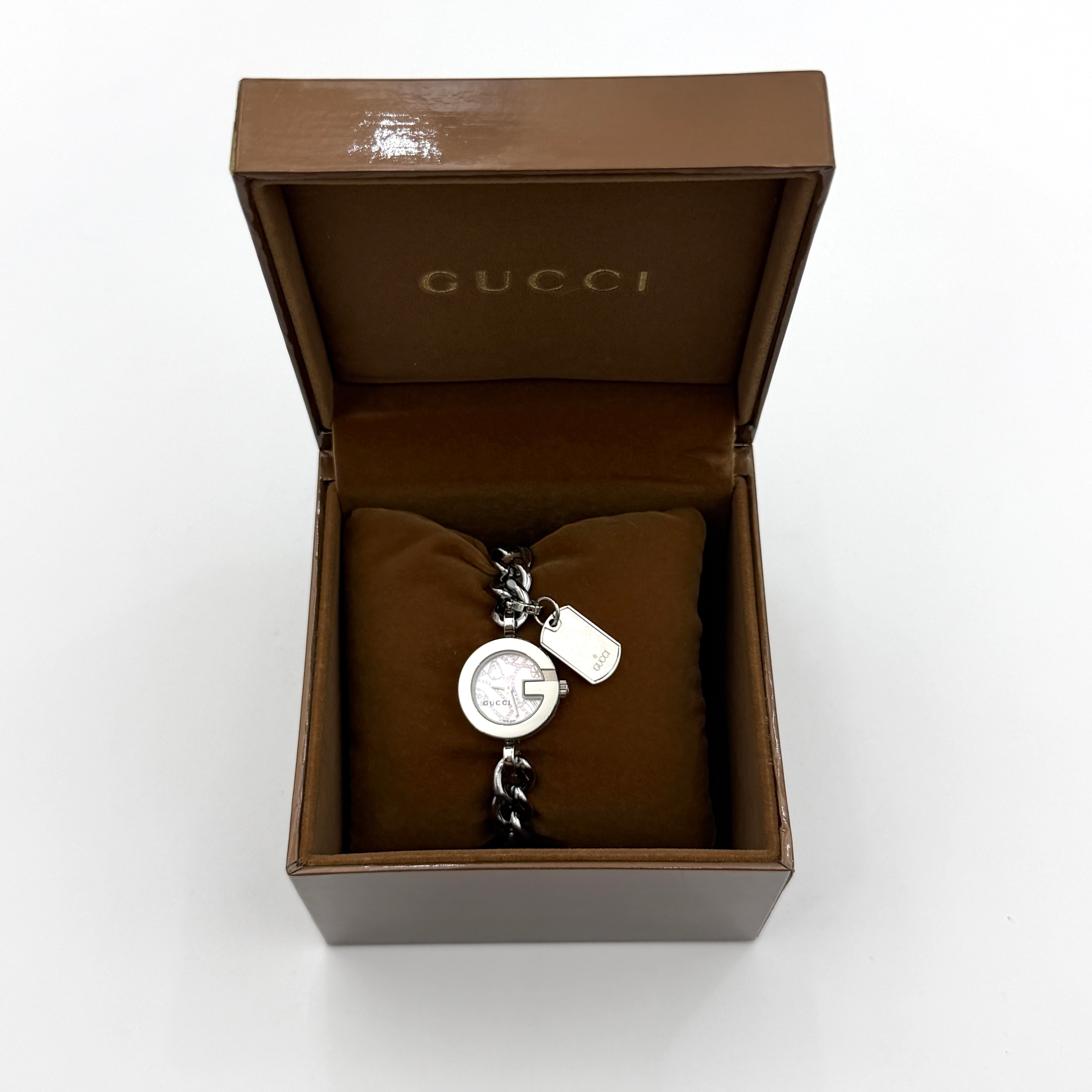 Gucci 107 G Charm Bracelet Watch with silver chunky chain and pink dial, styled on a brown cushion with original Gucci box.