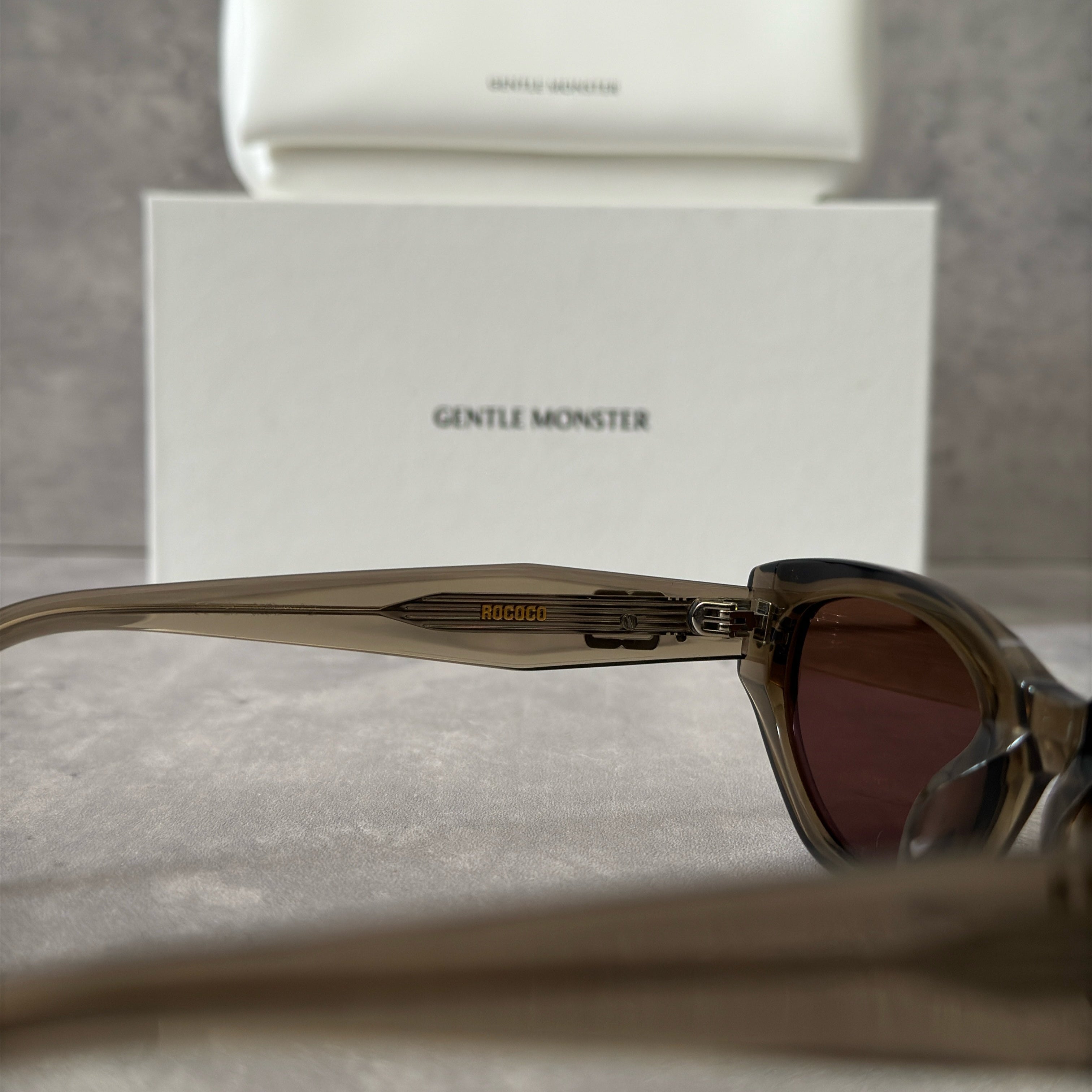 Gentle Monster ROCOCO KC6 sunglasses in translucent smoky brown acetate, featuring a bold sculpted design, tinted brown lenses, and signature metal temple detailing.