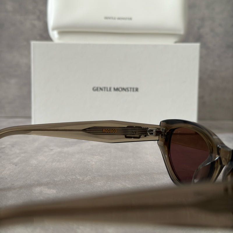 Gentle Monster ROCOCO KC6 sunglasses in translucent smoky brown acetate, featuring a bold sculpted design, tinted brown lenses, and signature metal temple detailing.
