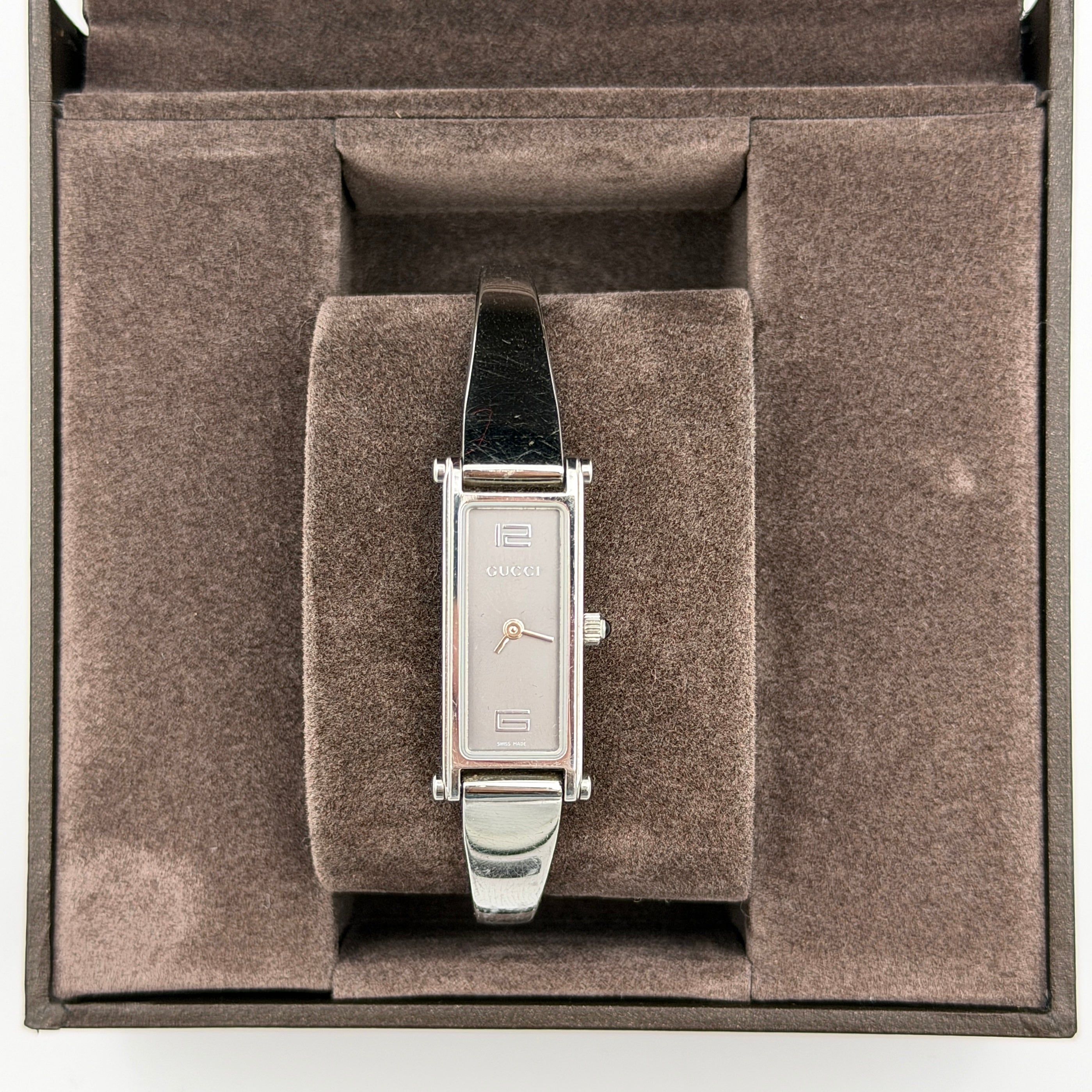 The Gucci 1500L Grey Dial Watch rests elegantly on its plush brown cushion atop a branded box, showcasing its sleek rectangular design and polished silver-tone bracelet.