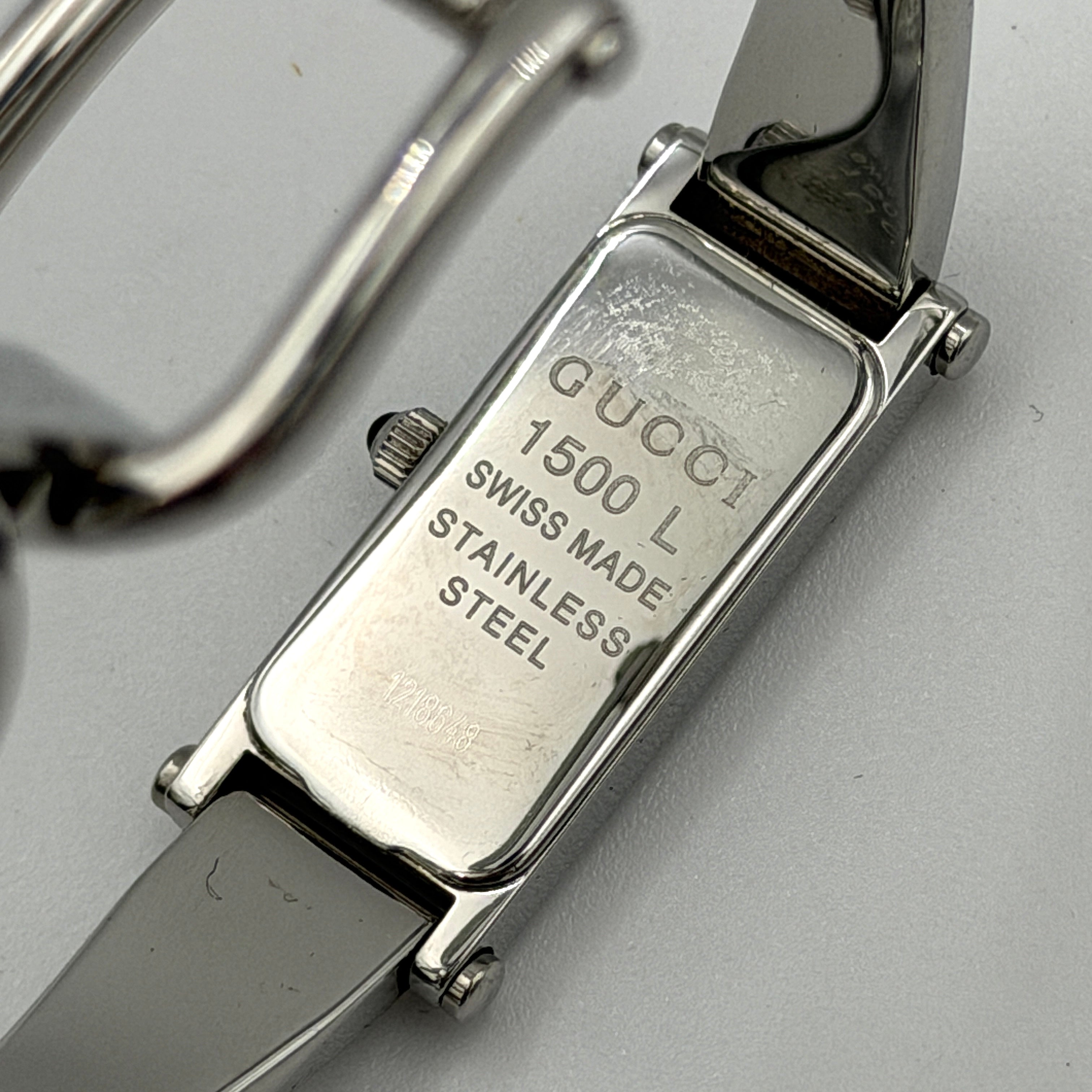 The Gucci 1500L Grey Dial Watch rests elegantly on its plush brown cushion atop a branded box, showcasing its sleek rectangular design and polished silver-tone bracelet.