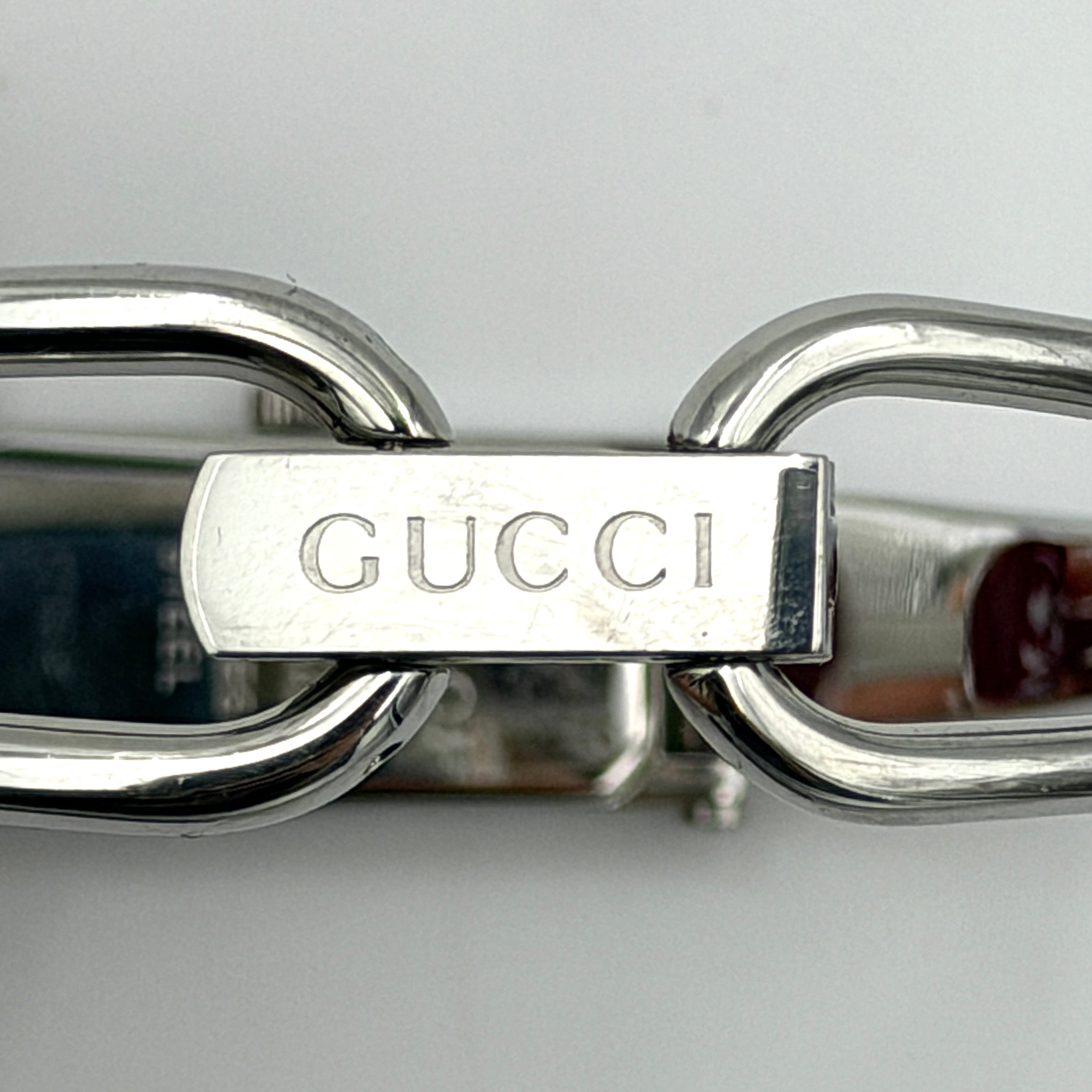 The Gucci 1500L Grey Dial Watch rests elegantly on its plush brown cushion atop a branded box, showcasing its sleek rectangular design and polished silver-tone bracelet.