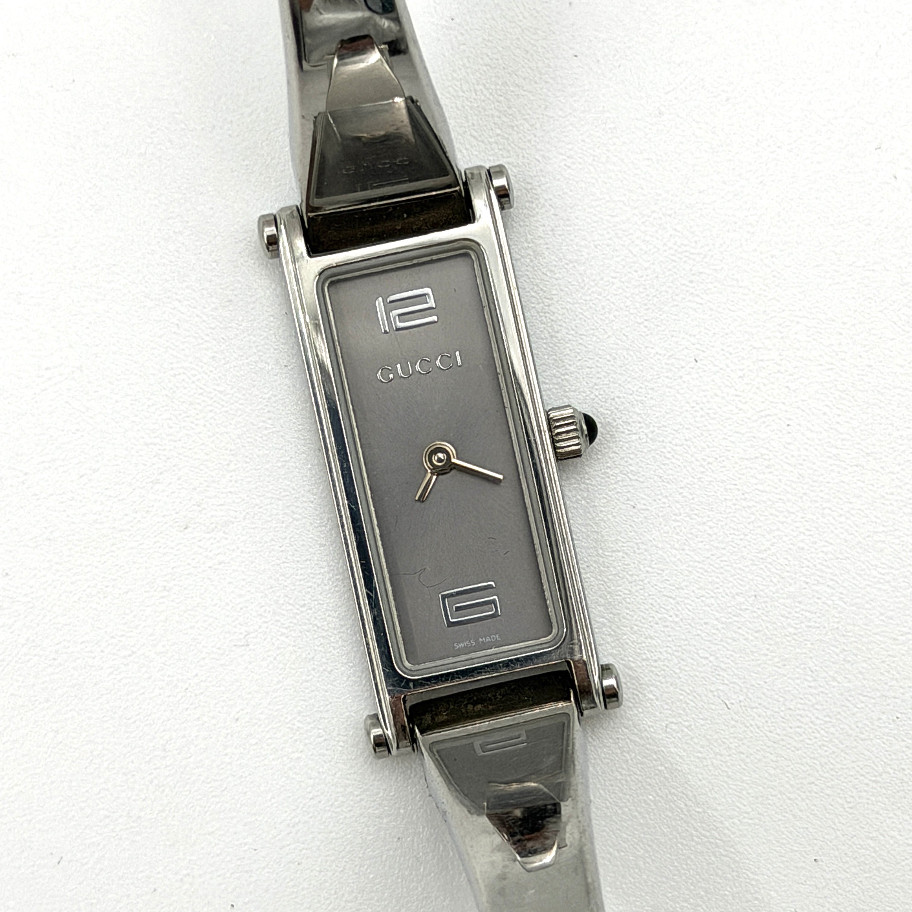 The Gucci 1500L Grey Dial Watch rests elegantly on its plush brown cushion atop a branded box, showcasing its sleek rectangular design and polished silver-tone bracelet.