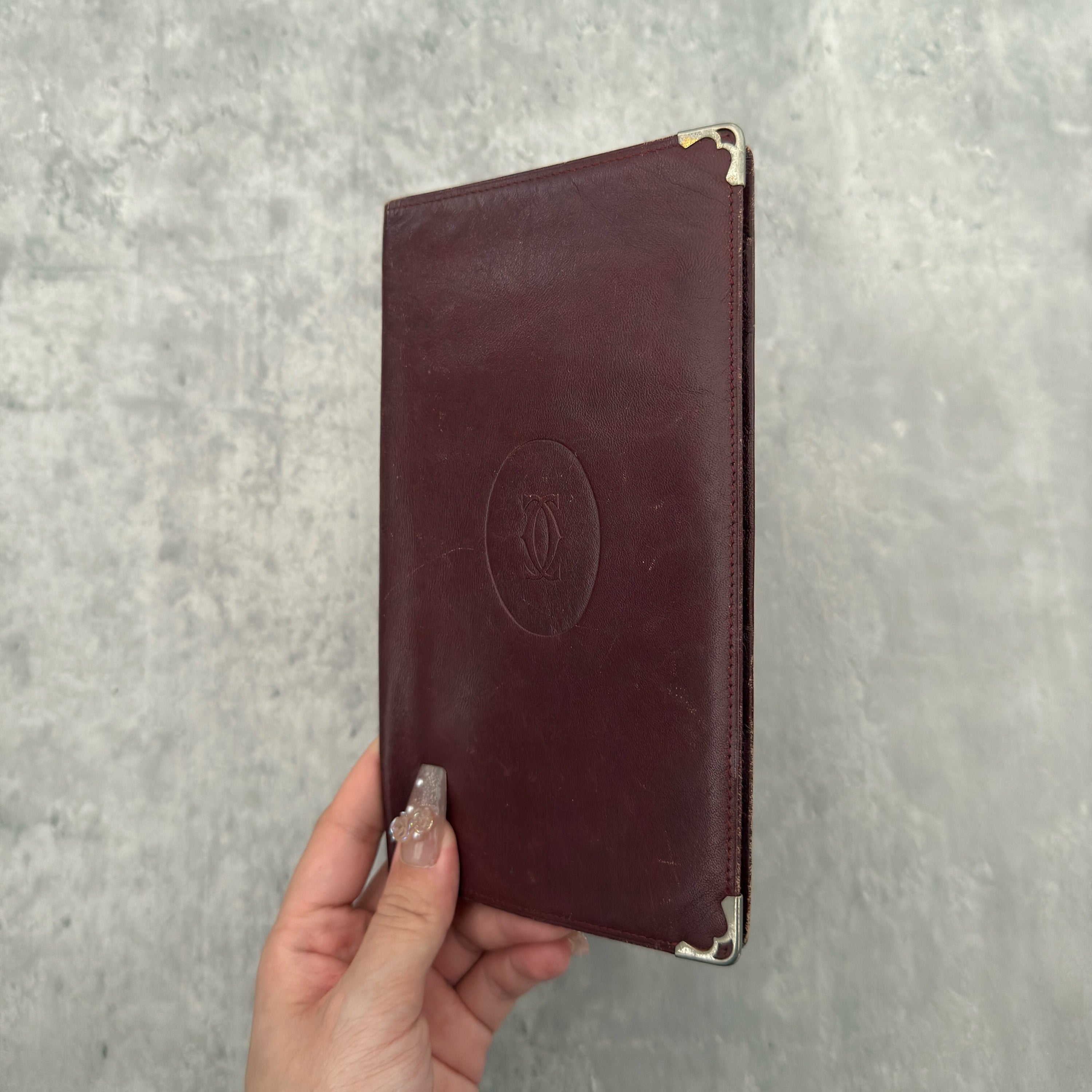 Cartier Must De Cartier Long Wallet in burgundy leather with embossed logo, gold-tone corner accents, and multiple compartments for organised storage.