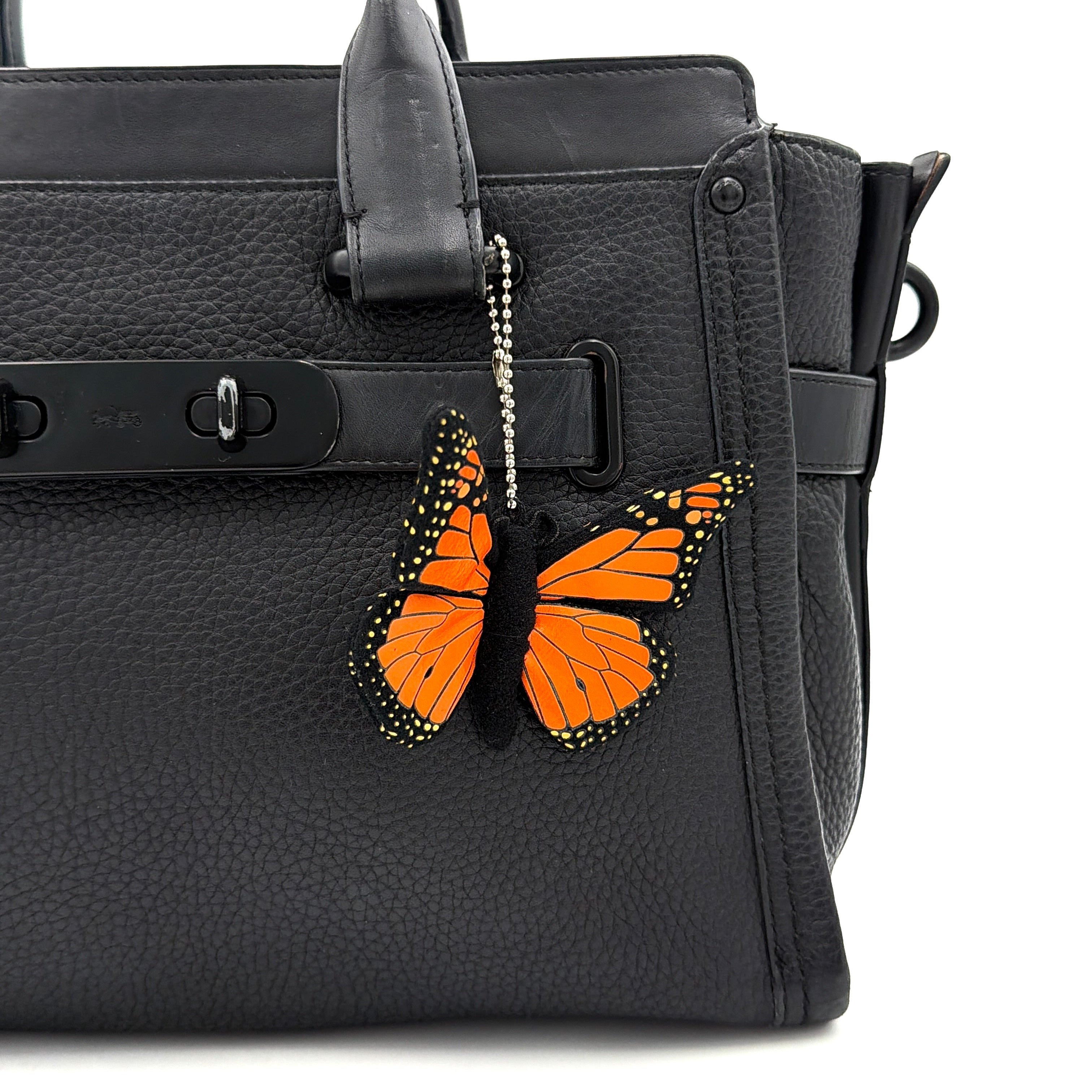 Coach Swagger Carryall handbag in black pebbled leather with matte black hardware and a vibrant orange butterfly charm.