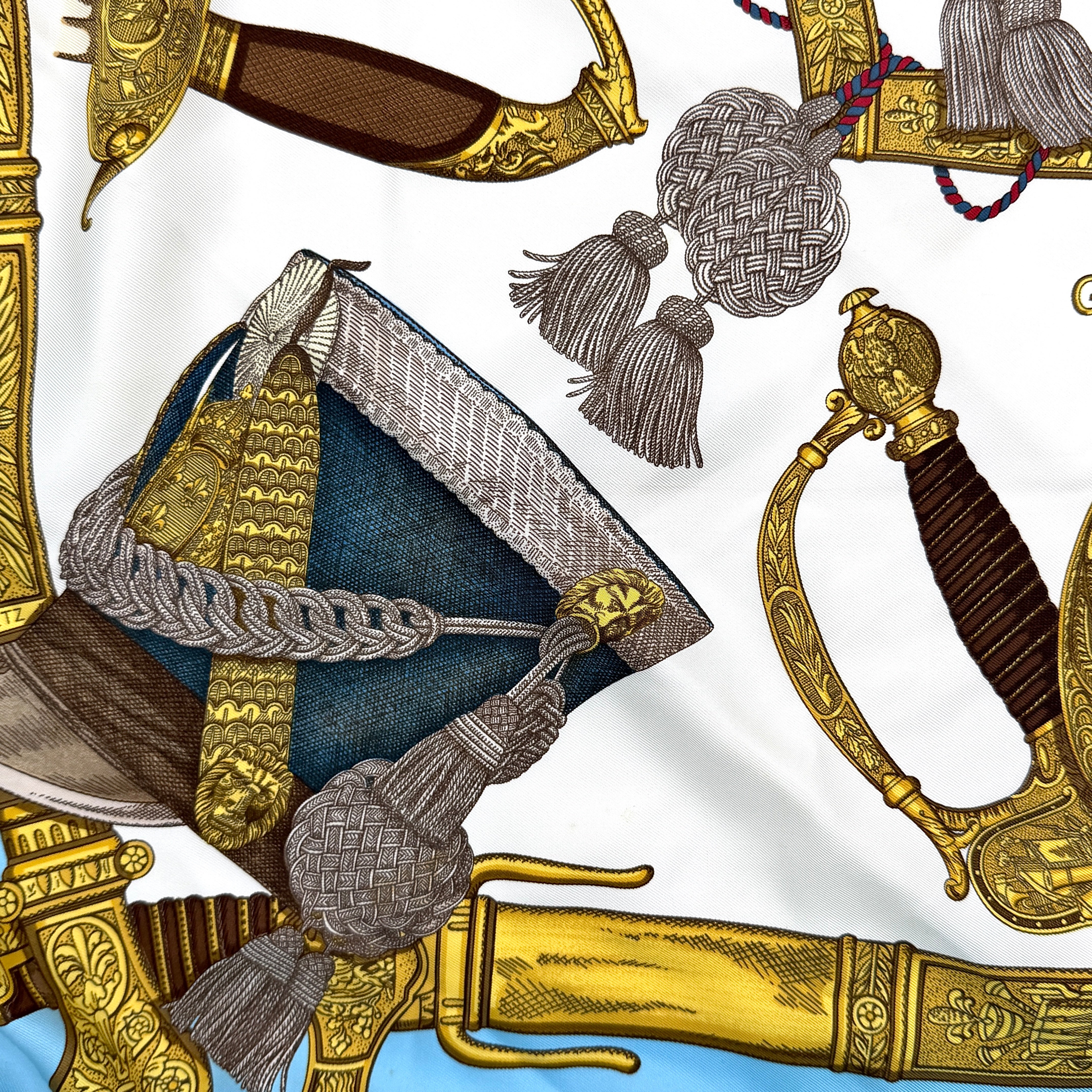 Hermès Grand Uniforme silk scarf with intricate gold and blue Napoleonic-inspired motifs framed on a soft blue and ivory background.